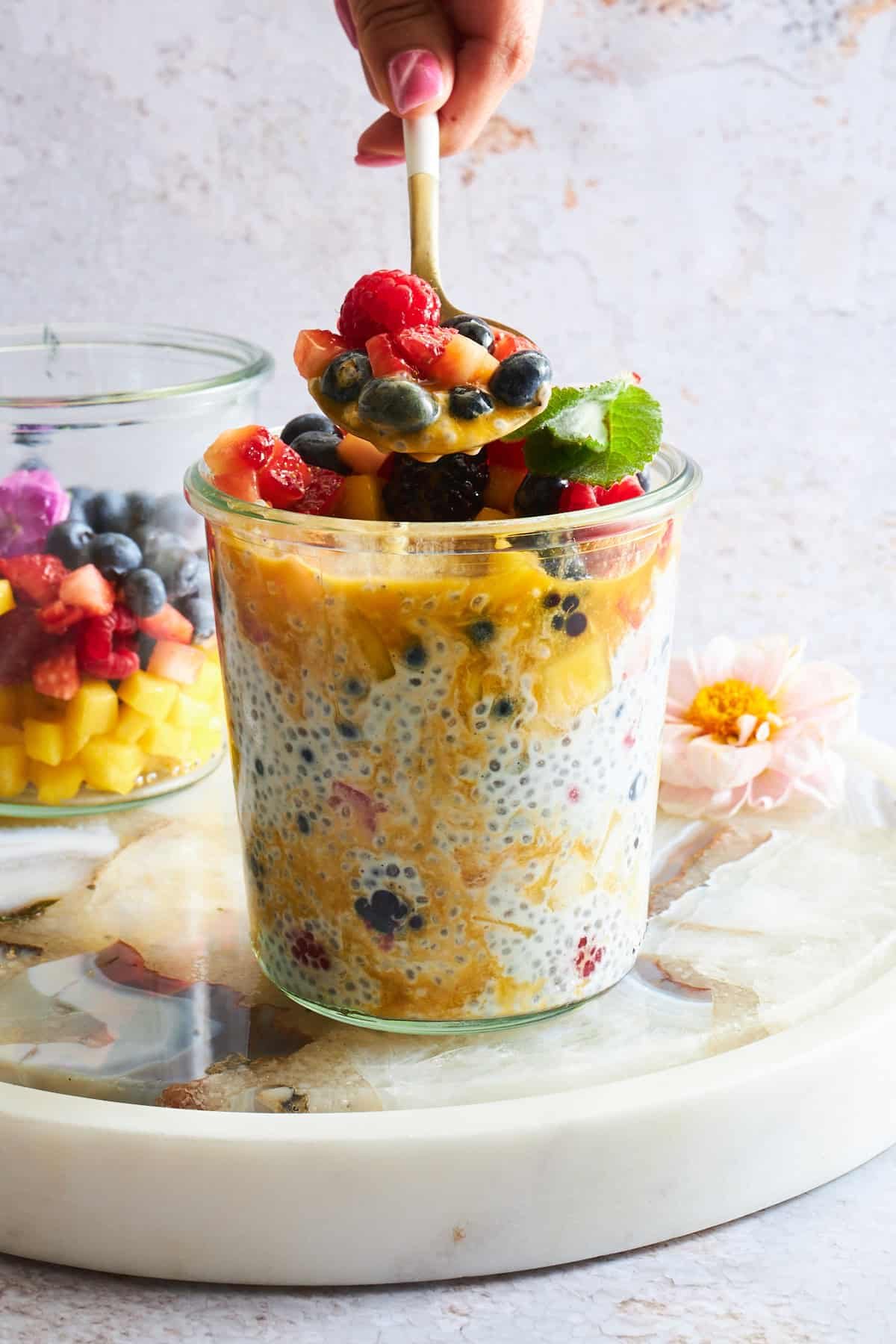 A jar of mango chia pudding topped with fruit salad with a spoon lifting a scoop from the top.