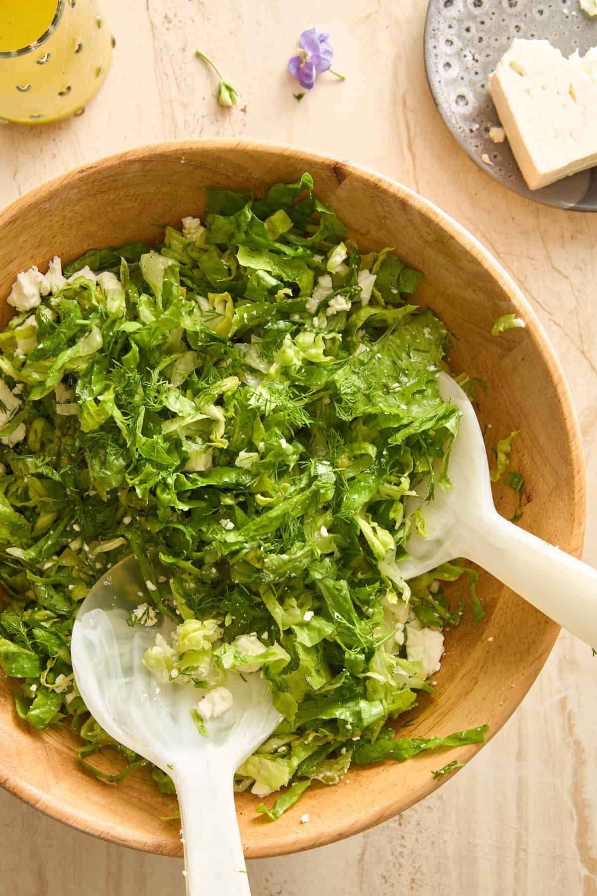 A wooden bowl filled with chopped green salad and feta cheese, with two white salad servers. Nearby are a plate with a block of cheese and a drink in a glass, set on a light marble surface. Ingredients for maroulosalata.