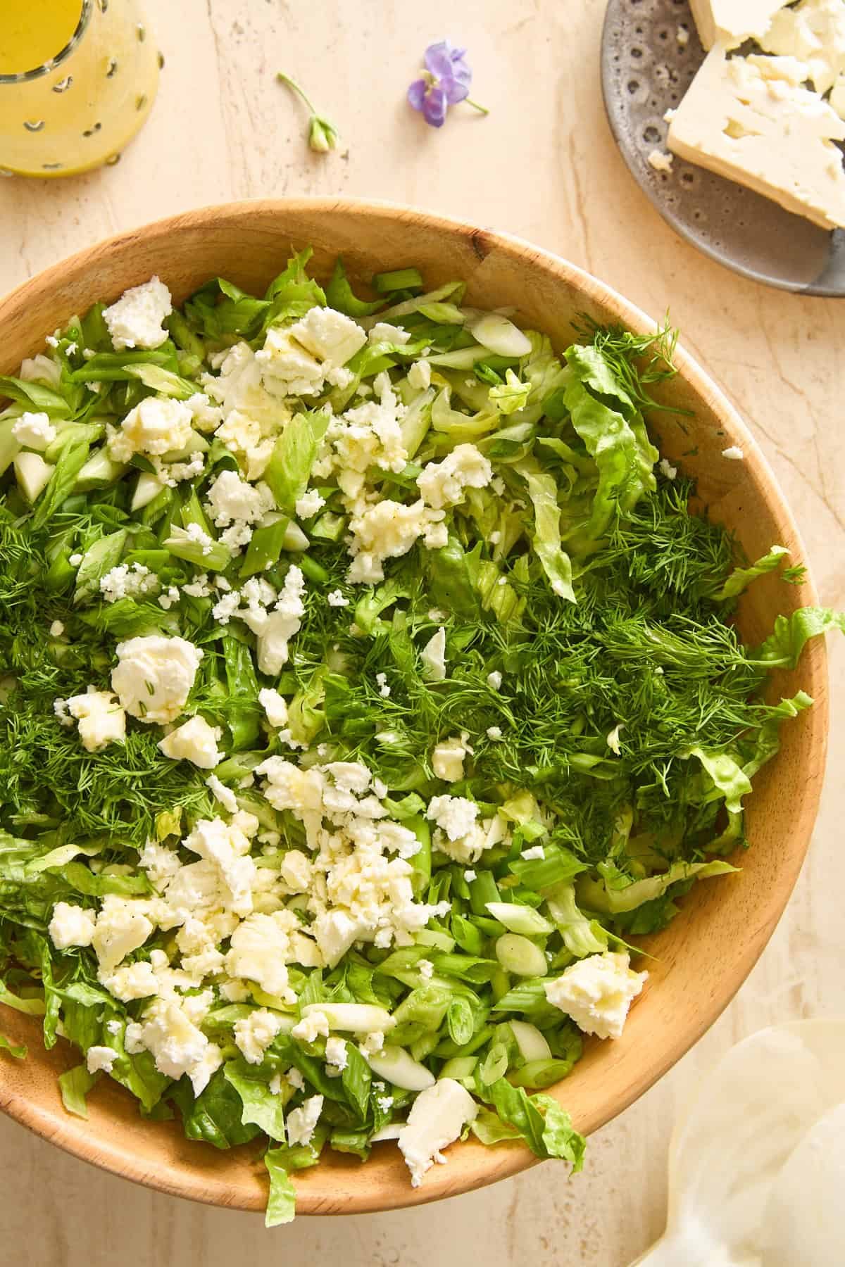 A wooden bowl filled with chopped lettuce, fresh dill, green onions, and crumbled feta cheese. A plate with a chunk of feta and a jar of dressing are nearby on a light countertop. Ingredients for maroulosalata.