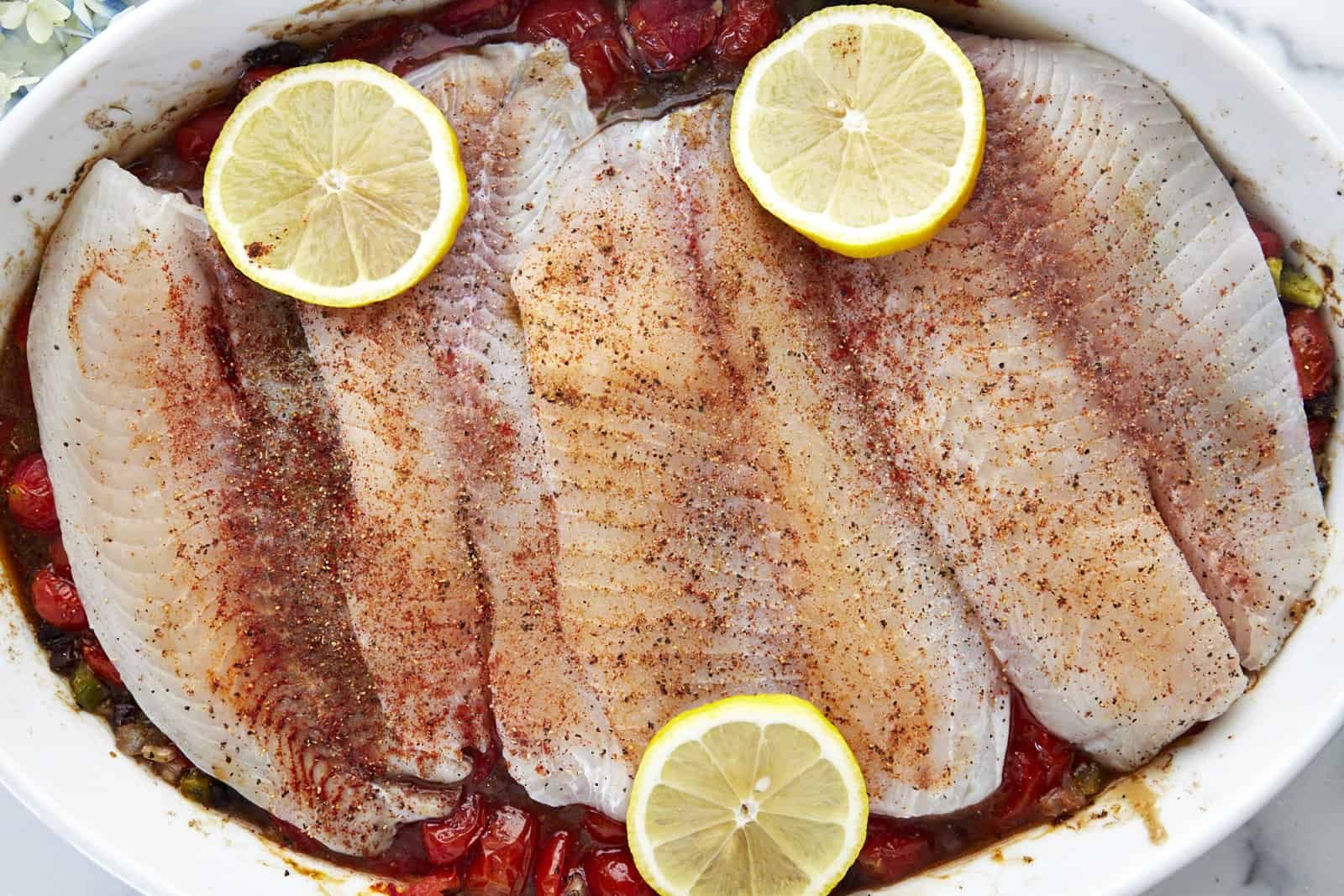 Four raw tilapia filets over cooked a tomato mixture topped with lemon slices.