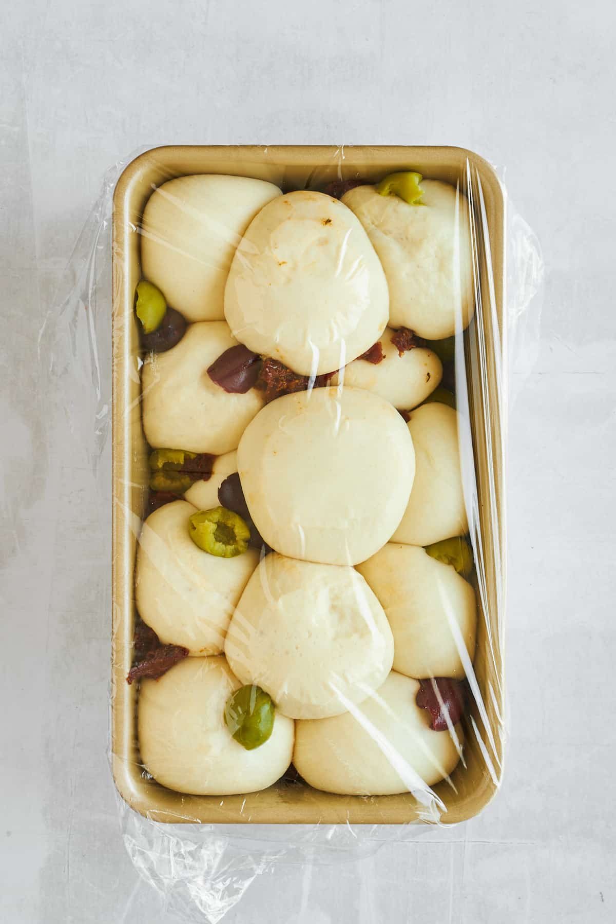 Raw bread rolls, olives, and sun-dried tomatoes in a loaf pan covered with plastic wrap.