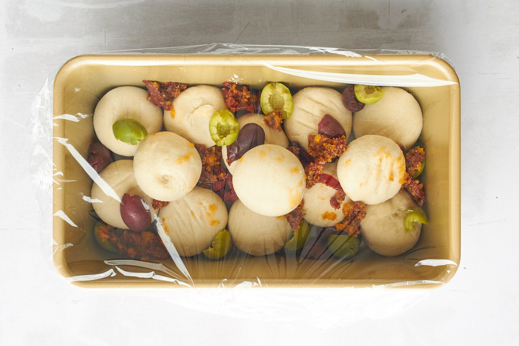 Raw dinner rolls topped with kalamata olives and sun-dried tomatoes covered with plastic wrap.