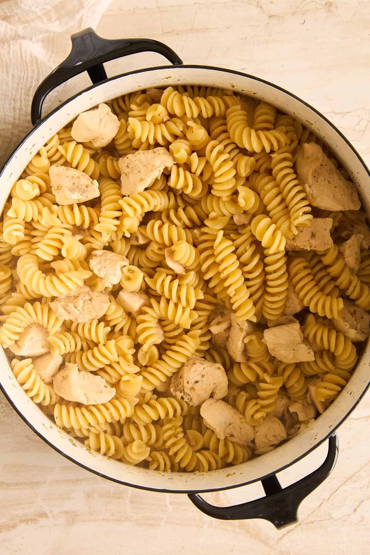 A large white pot filled with cooked rotini pasta and pieces of seasoned chicken sits on a light-colored surface.