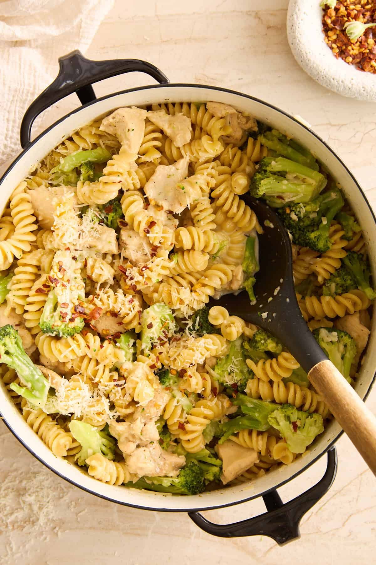 A pot of rotini pasta mixed with pieces of chicken and broccoli, topped with grated cheese, sits on a light countertop. A black serving spoon rests in the pot, and a bowl of red pepper flakes is nearby.