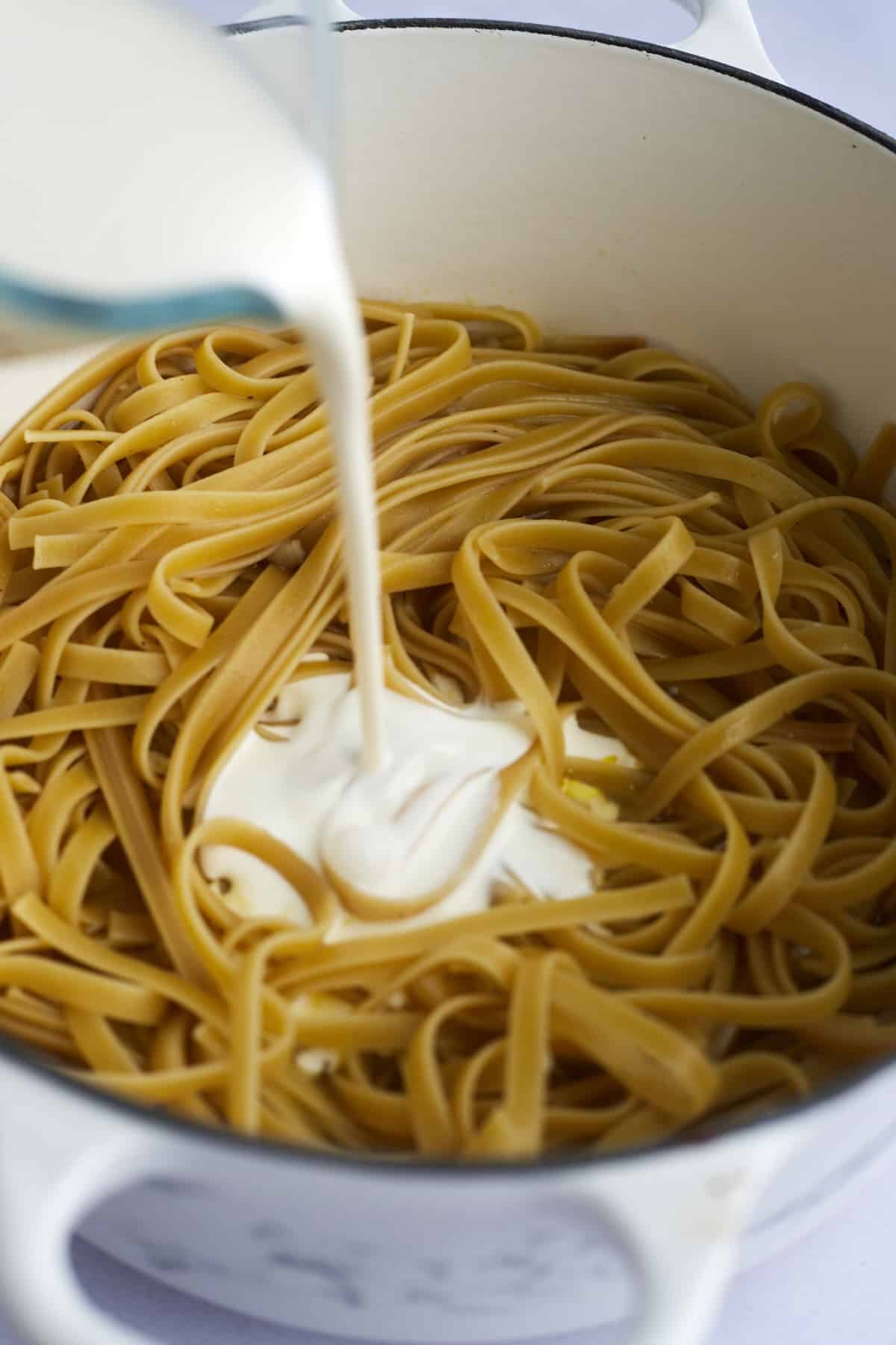 Alfredo sauce being poured over cooked fettuccine noodles in a pot.