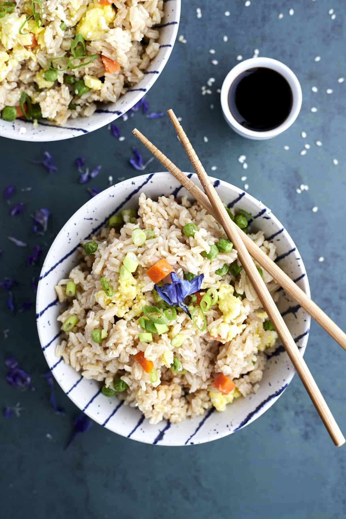 A bowl of simple fried rice with veggies with two chopsticks balanced on top and a side of soy sauce