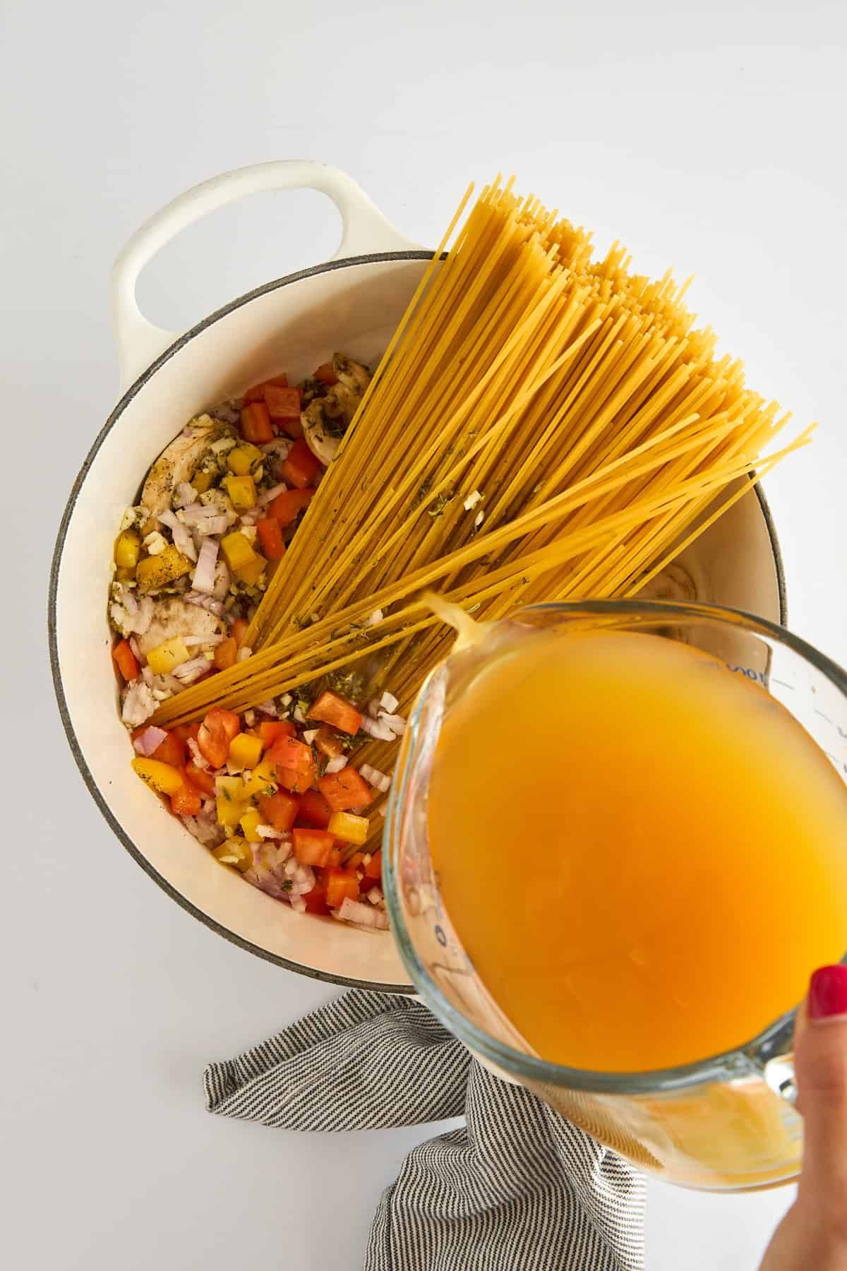 Broth being poured into a pot of sautéed veggies and raw spaghetti noodles.