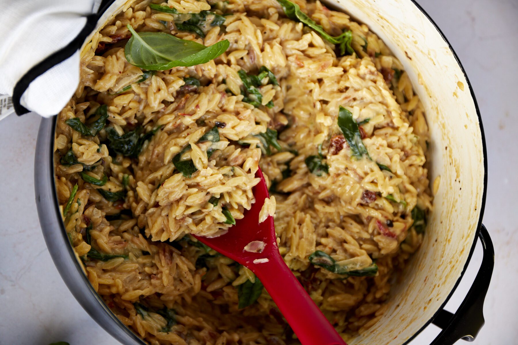 Creamy Tuscan orzo in a pot, a red spoon lifting a scoop.