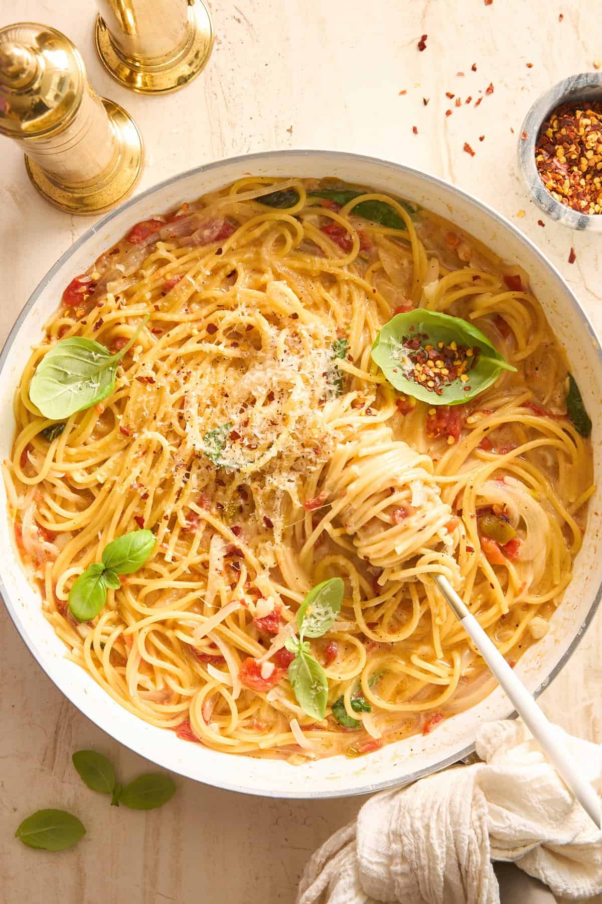 A skillet filled with silky spaghetti garnished with fresh basil and grated cheese. A fork twirls some pasta, and there are gold salt and pepper shakers, red pepper flakes, and basil leaves nearby on a light table.