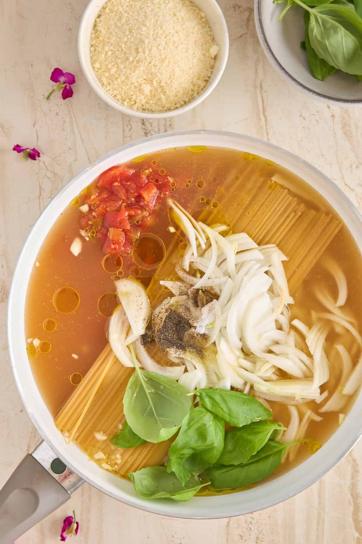 A pot filled with uncooked spaghetti, sliced onions, chopped tomatoes, garlic, basil leaves, broth, oil, and spices sits on a light countertop; nearby are bowls of grated cheese and fresh basil leaves.
