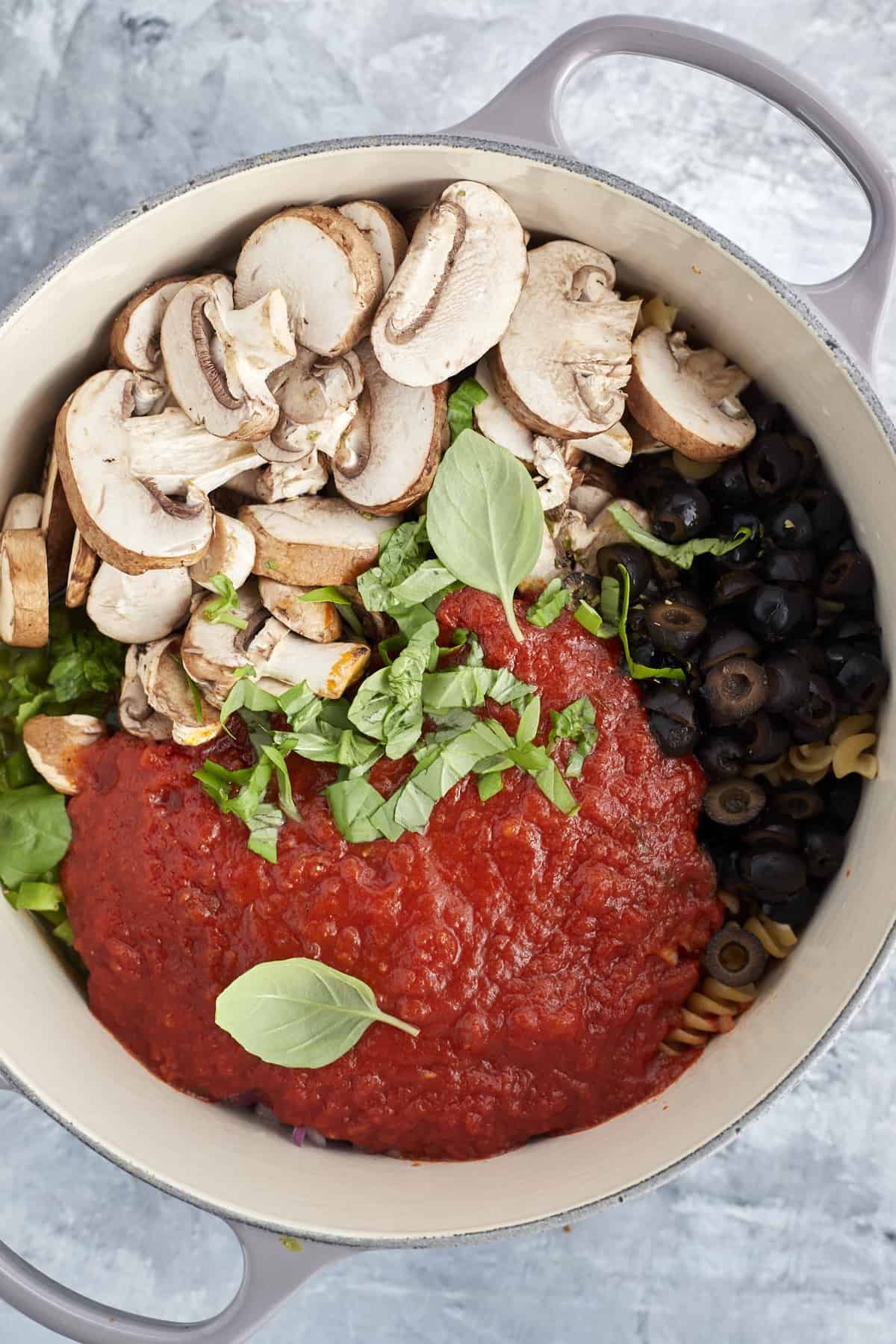 overhead image of a pot full of uncooked pasta, marinara sauce, fresh herbs, mushrooms, and black olives