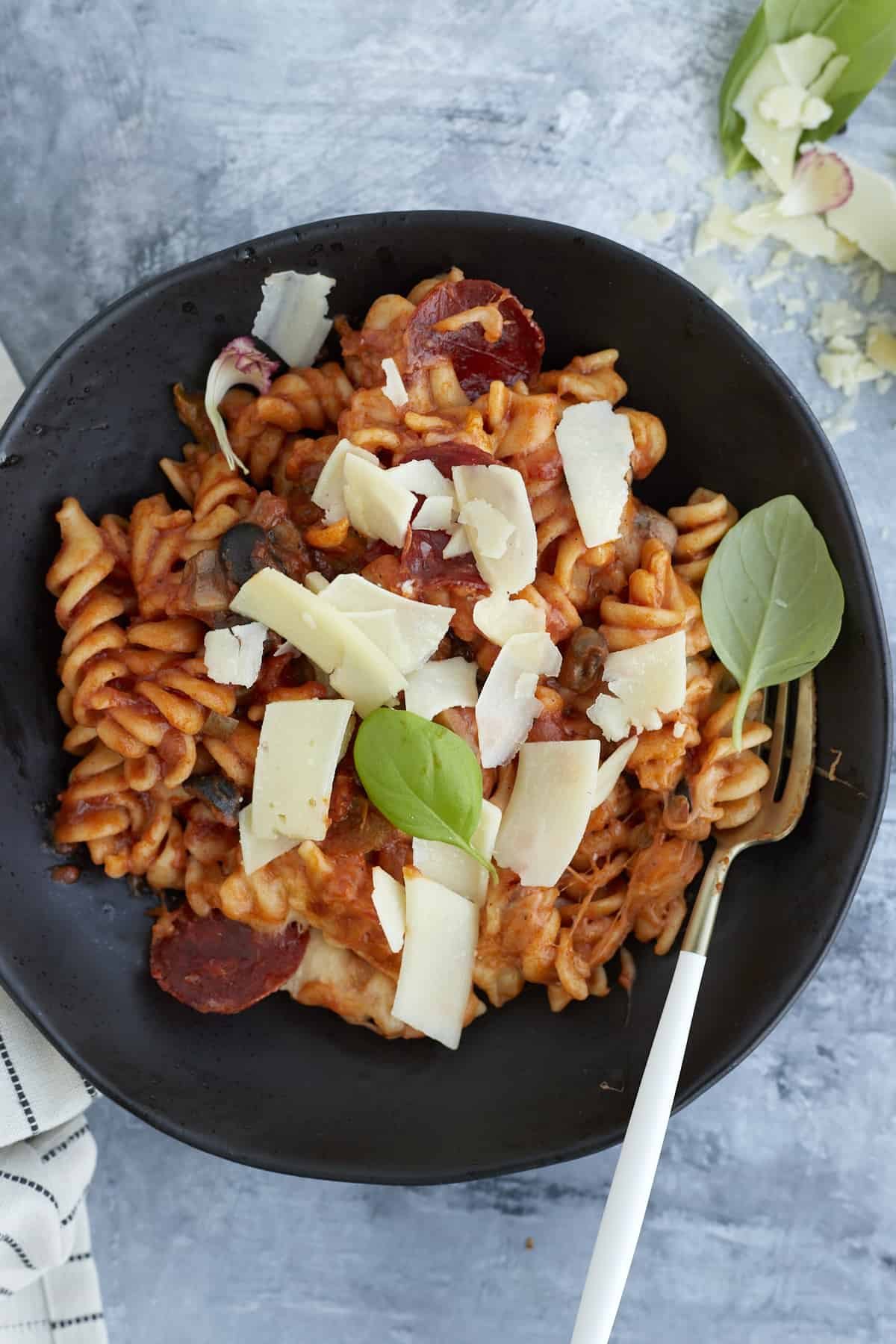overhead image of pizza pasta in a black bowl topped with shaved cheese and fresh herbs with a fork on the side