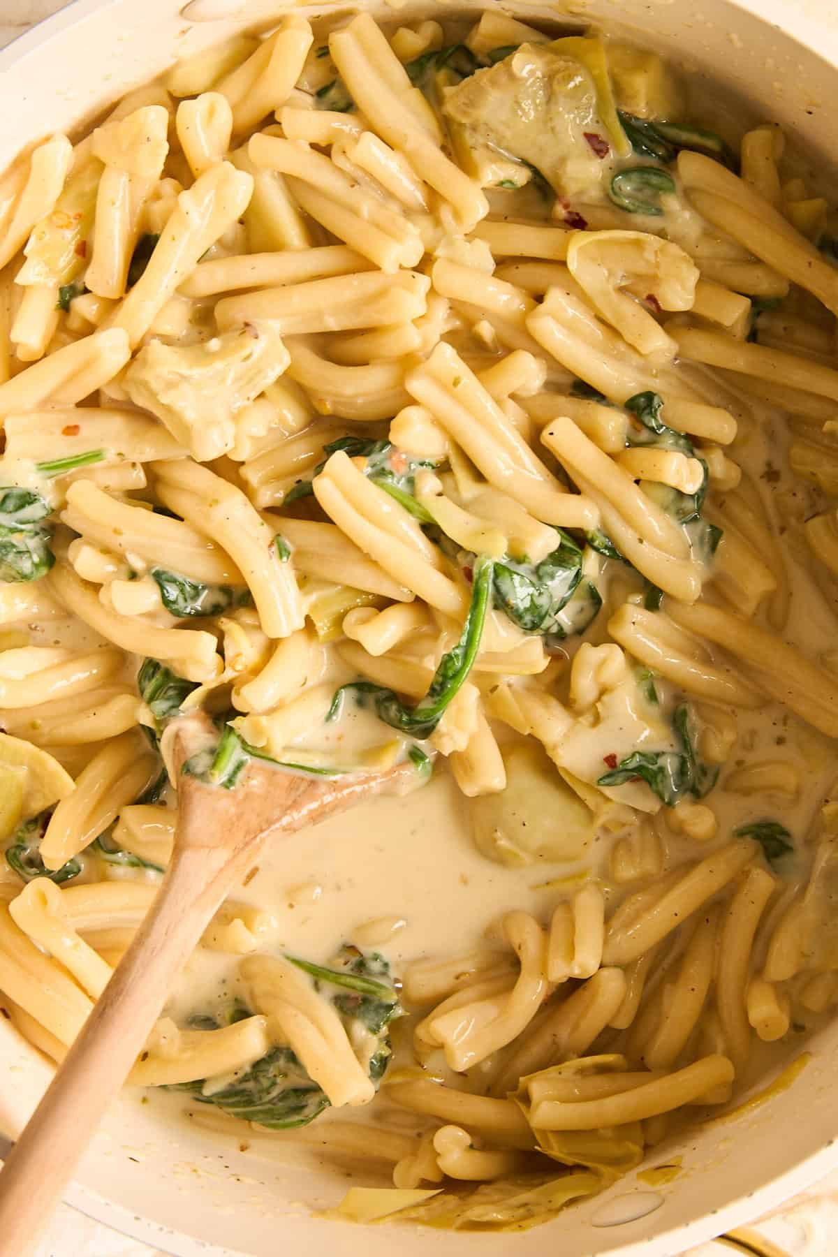 A pot of creamy pasta with spinach and artichoke pieces, being stirred with a wooden spoon. The sauce is rich and smooth, coating the pasta evenly.