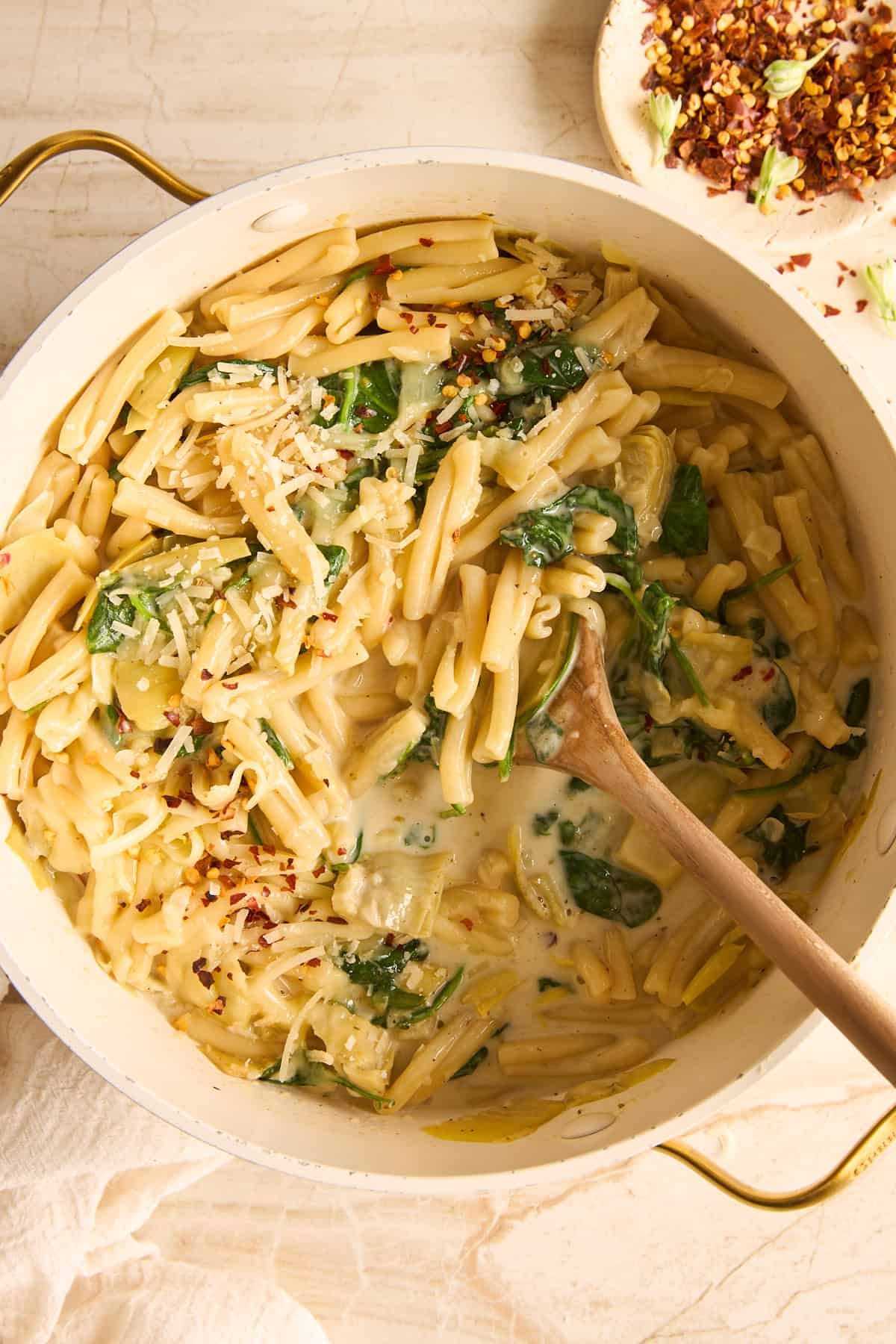A pot of silky pasta with spinach and artichokes, topped with grated cheese. A wooden spoon rests in the pasta, and a plate with red pepper flakes is visible in the background.