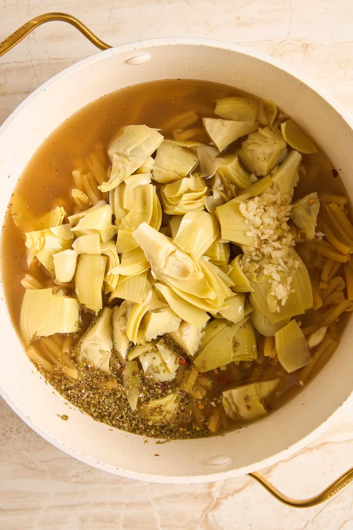A white pot filled with broth, uncooked pasta, chopped artichoke hearts, minced garlic, and dried herbs sits on a light-colored surface.