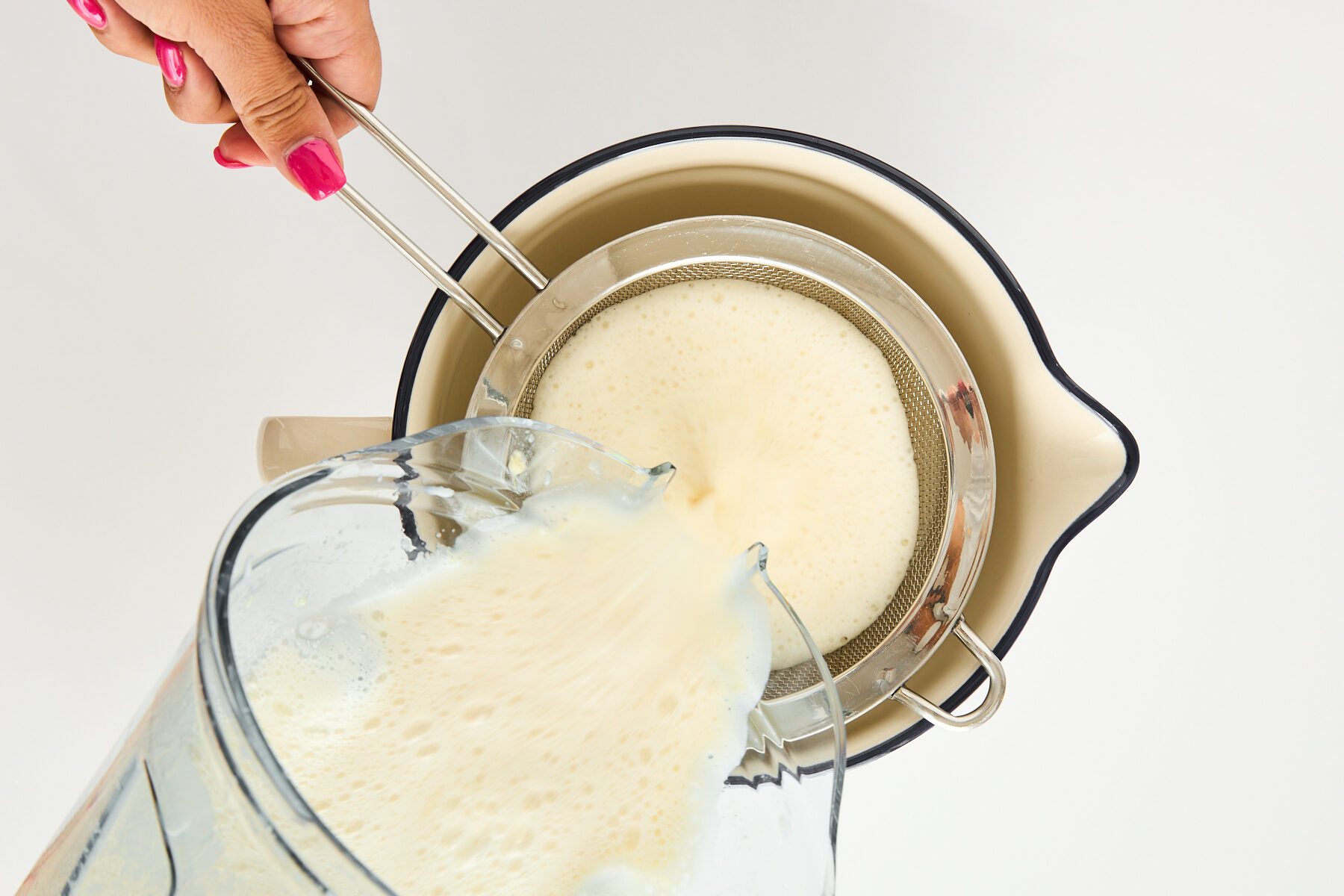 A hand holds a metal strainer over a cream-colored bowl while pouring a frothy liquid from a blender through the strainer. The person has pink nail polish. The background is white.