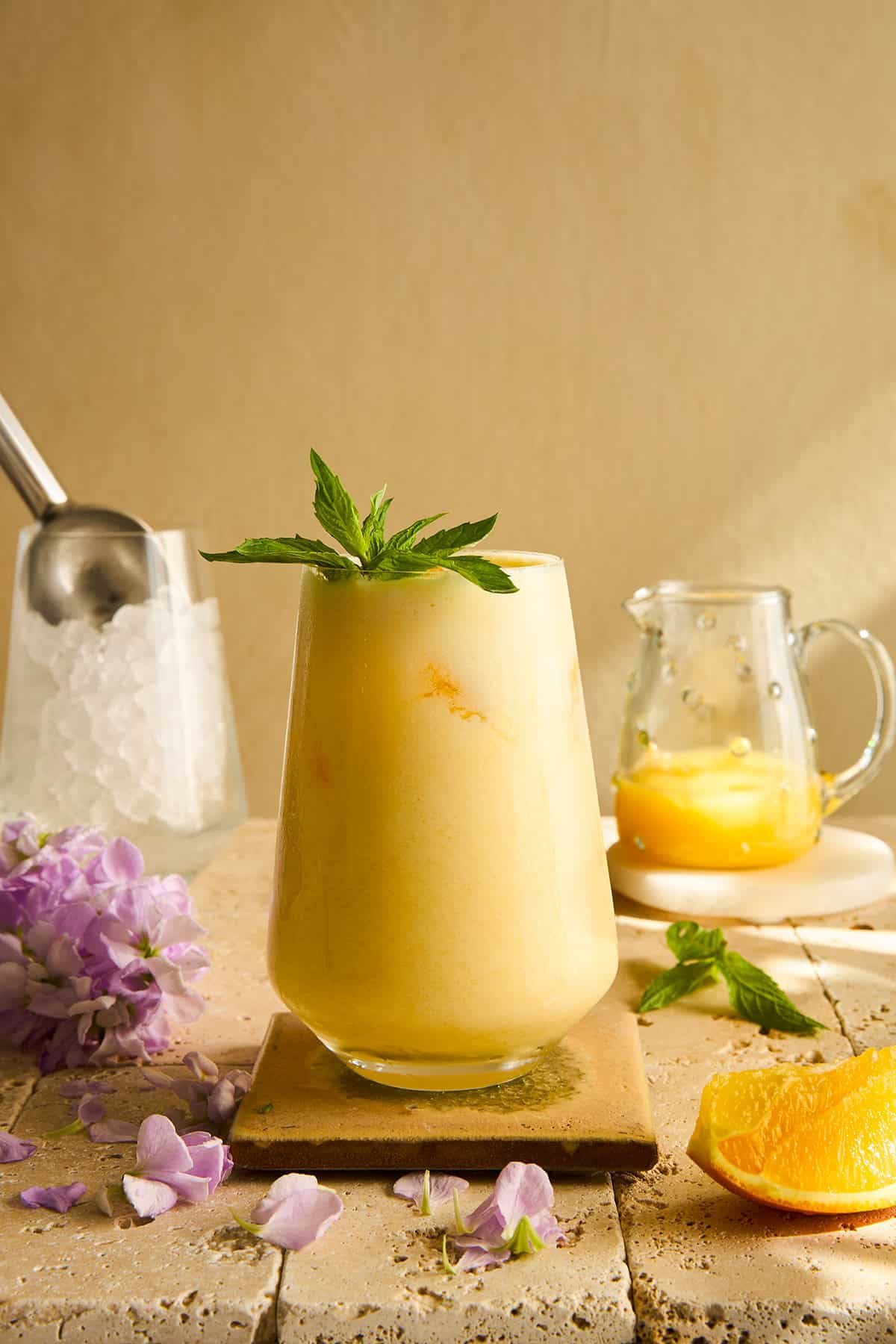 A silky orange smoothie garnished with a sprig of mint sits on a tile coaster, with a small pitcher of orange juice, an ice scoop, purple flowers, and an orange wedge nearby.