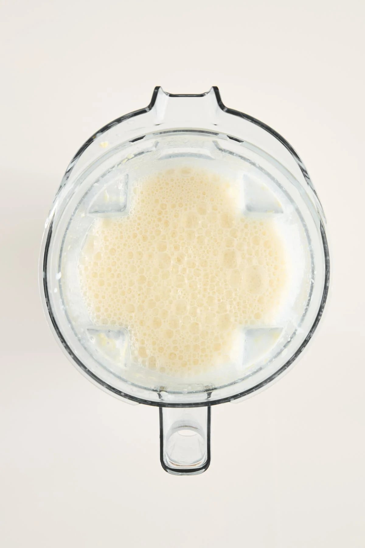 A top-down view of a blender filled with a light beige, frothy liquid, likely a smoothie or blended beverage, against a plain light background.