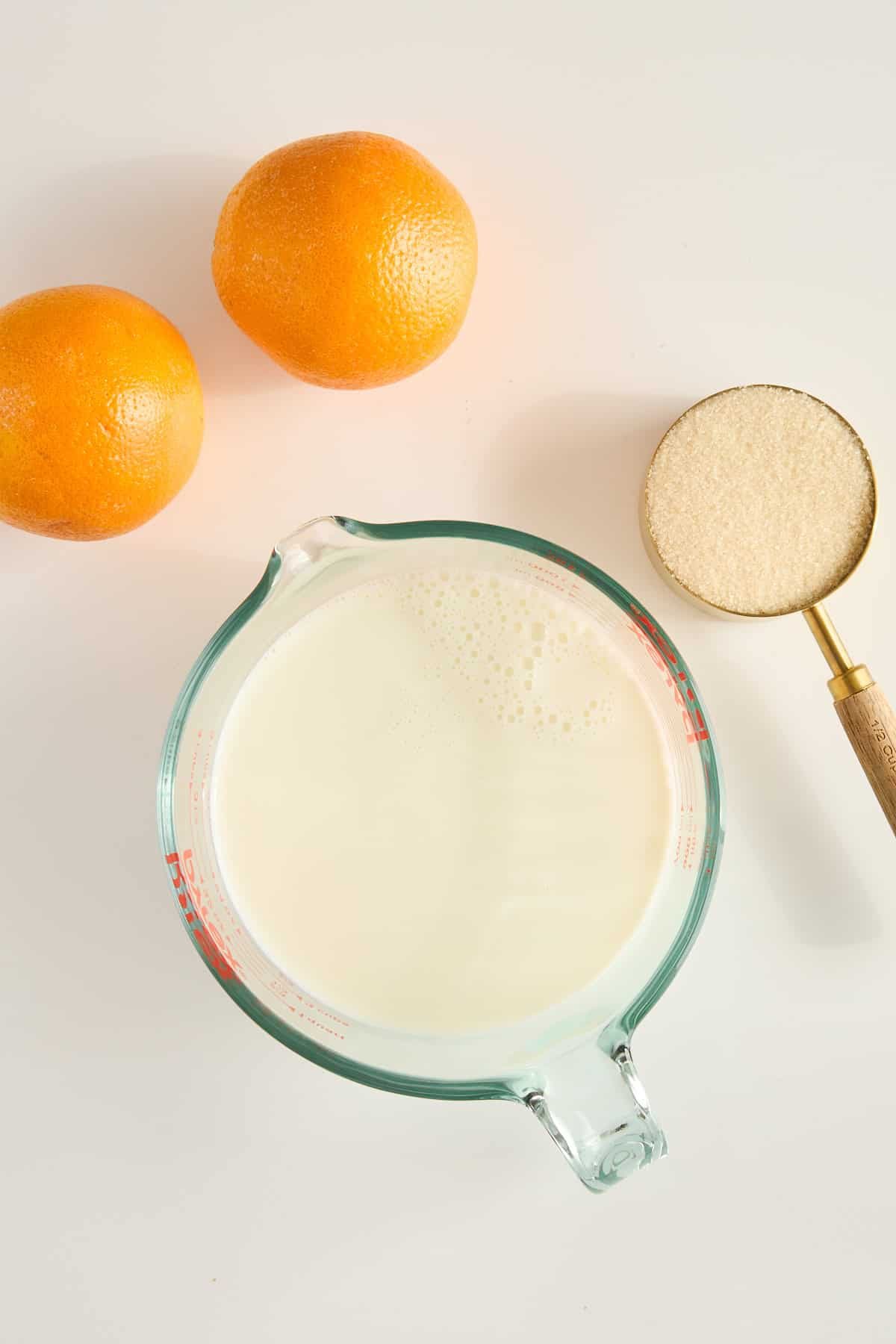 Two oranges, a glass measuring cup filled with milk, and a metal measuring cup with granulated sugar are arranged on a white surface.