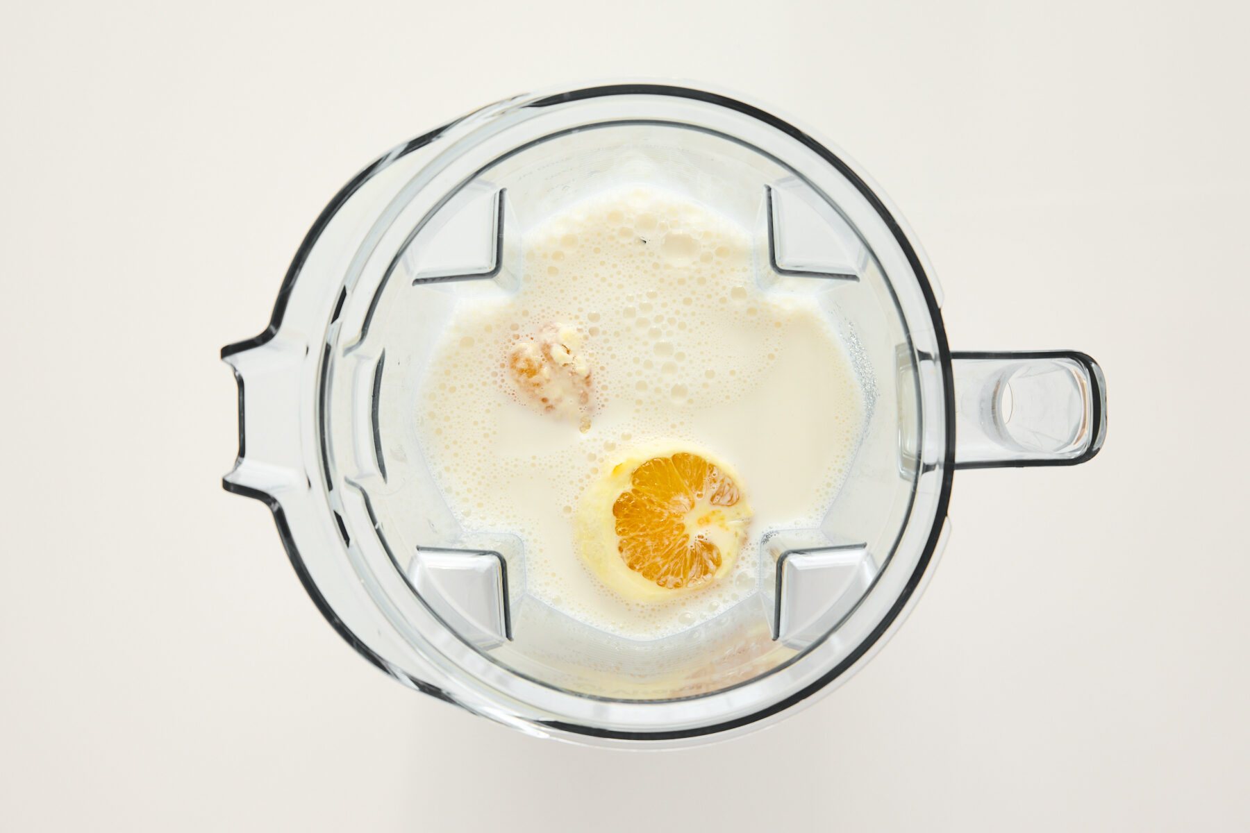 Top-down view of a blender containing milk, an orange half, and other ingredients, ready to be blended, on a white background.