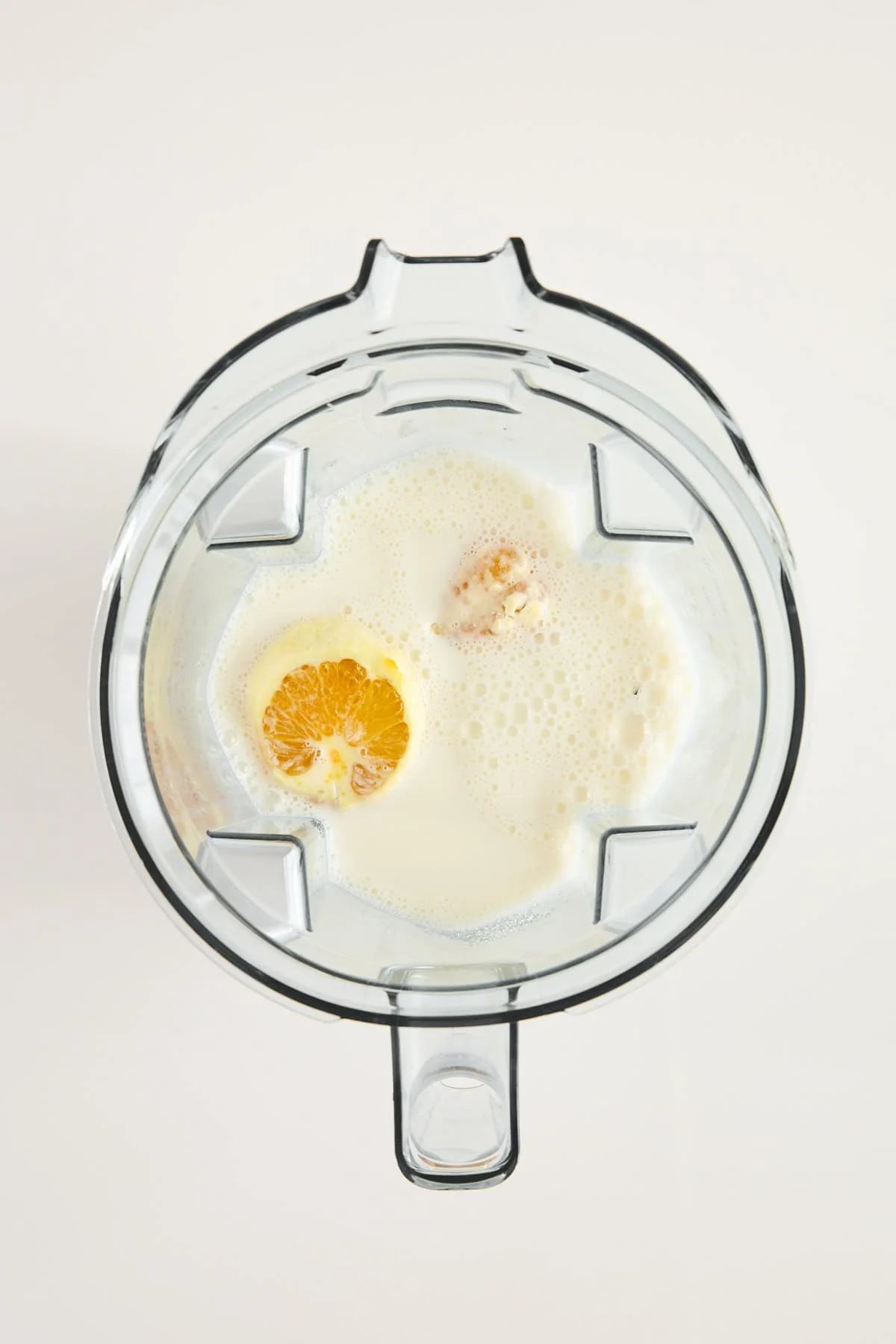 Top-down view of a blender containing milk, an orange half, and other ingredients, ready to be blended, on a white background.