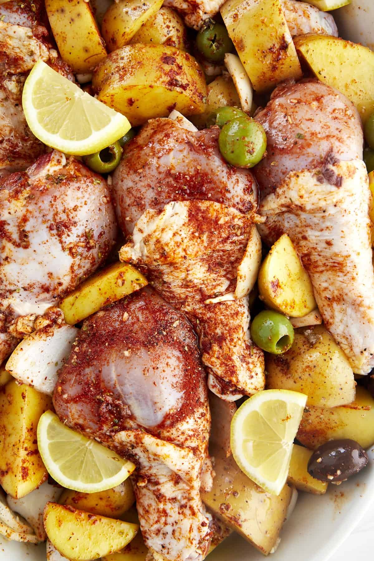 Raw chicken with golden potatoes, red and green olives, onions, and garlic coated with seasonings and topped with lemon wedges.