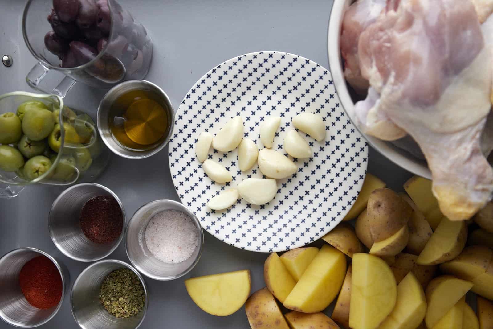 Raw chicken, golden potatoes, garlic cloves, spices, olive oil, green olives, and kalamata olives.