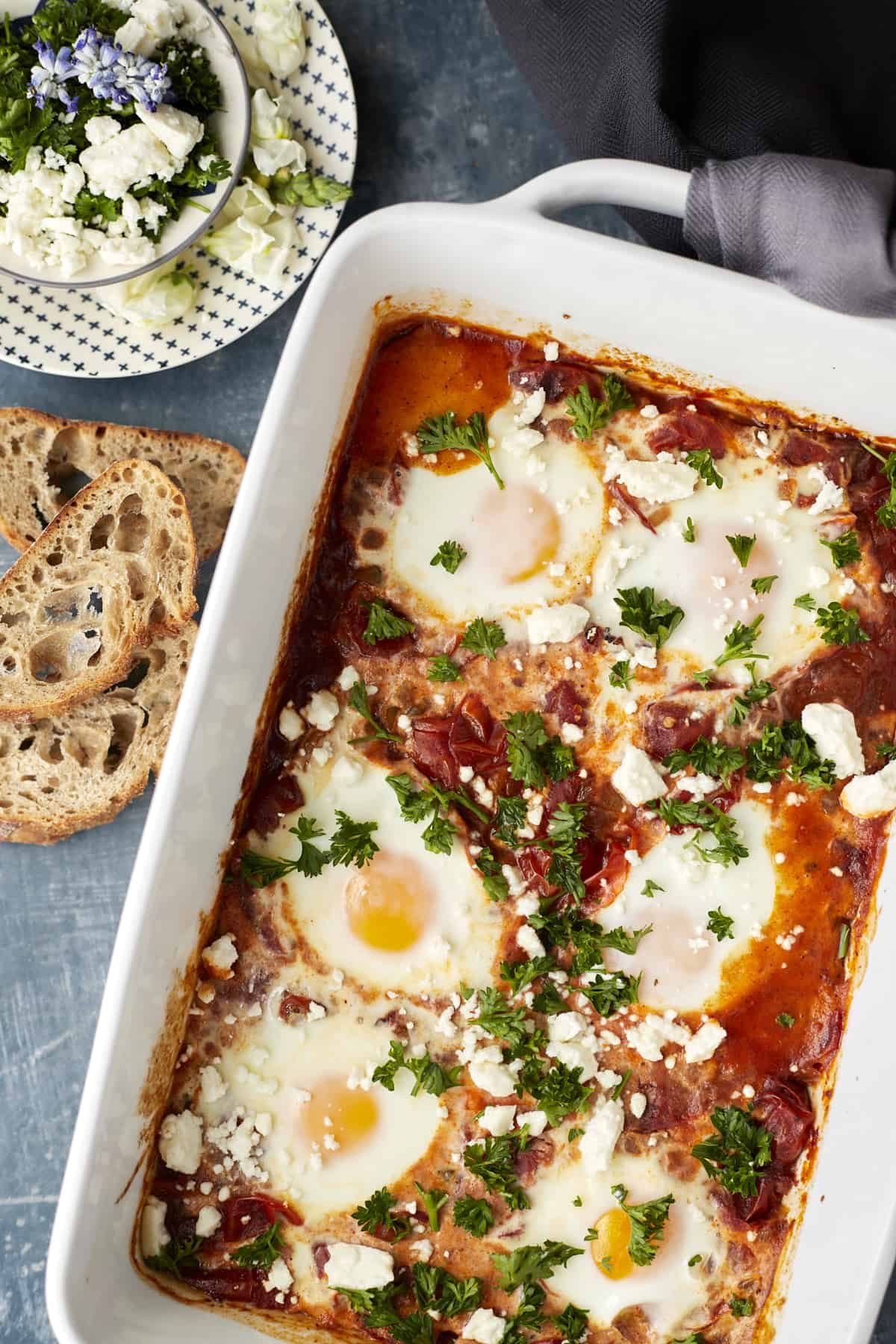 overhead image of oven baked shakshuka topped with herbs with pieces of bread to the left