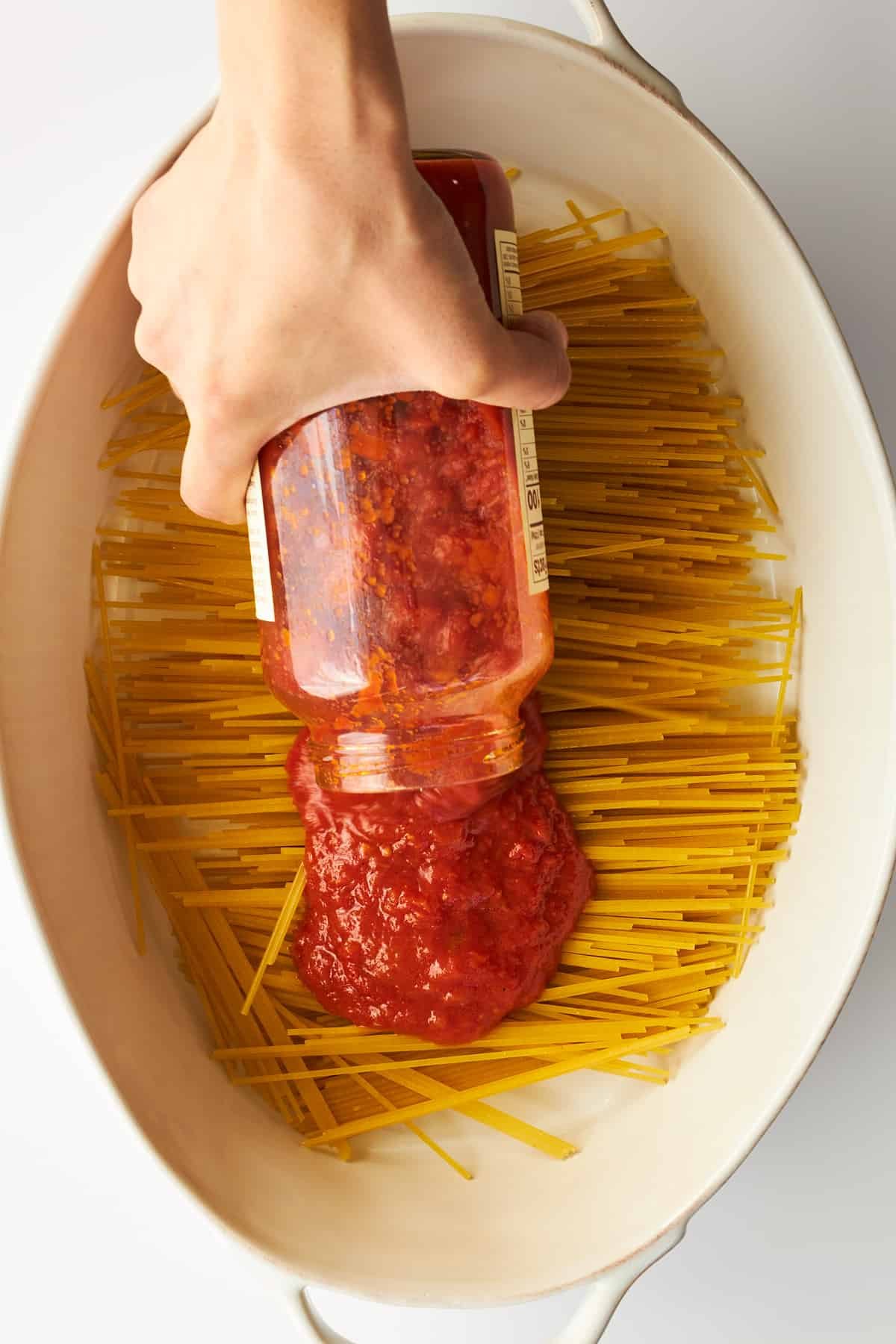 Marinara sauce being poured over spaghetti noodles in an oval baking dish.