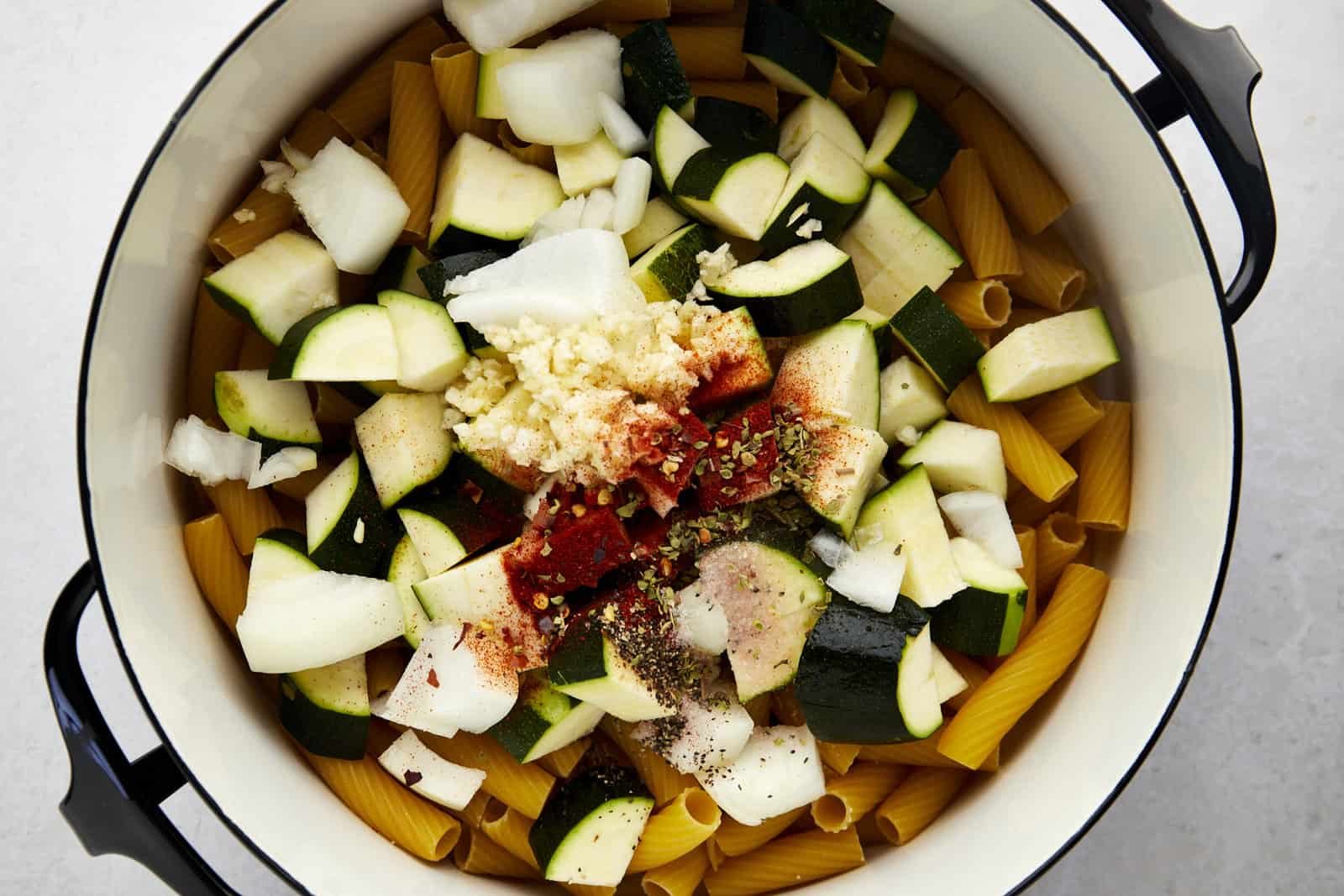 A pot full of rigatoni noodles, zucchini, onion, and spices.