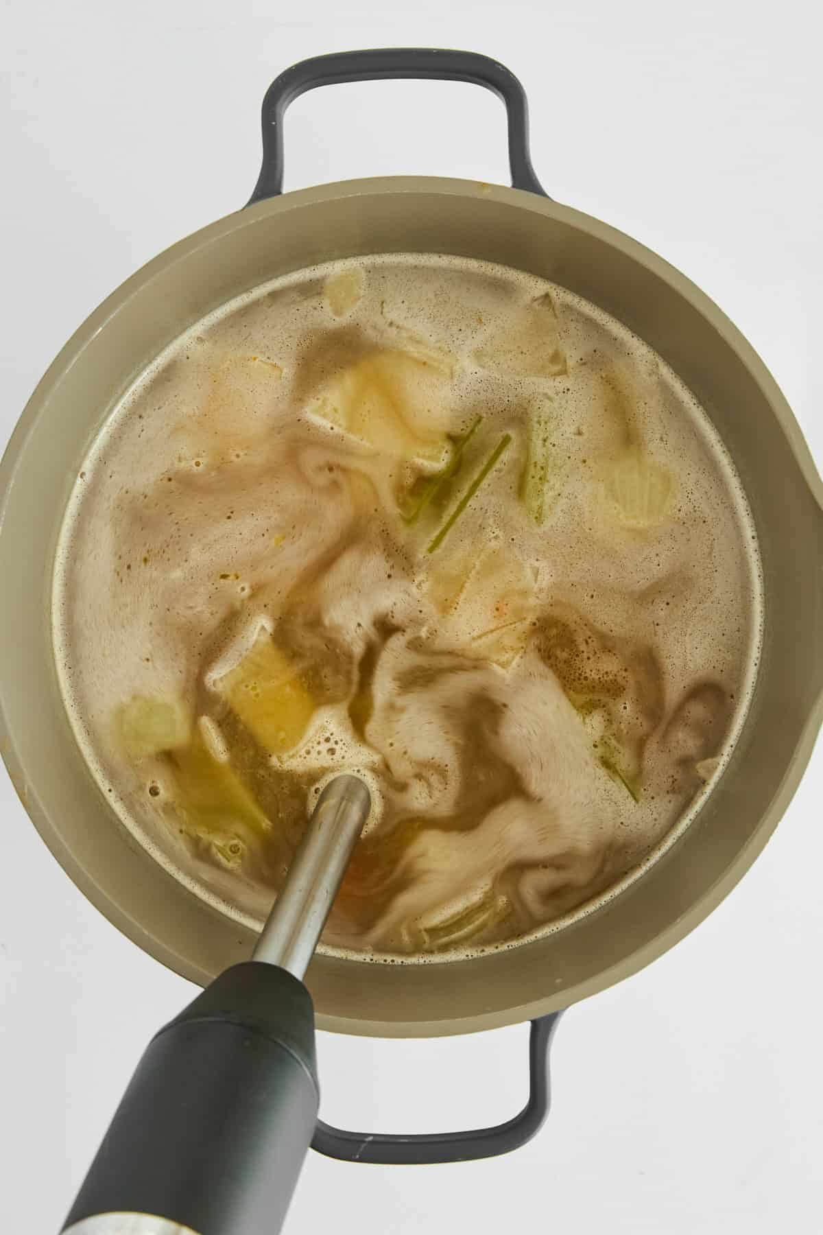 An immersion blender blending veggies into broth in a pot.