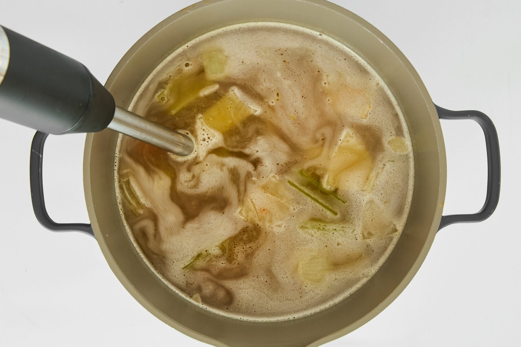 An immersion blender blending veggies in a pot of broth.