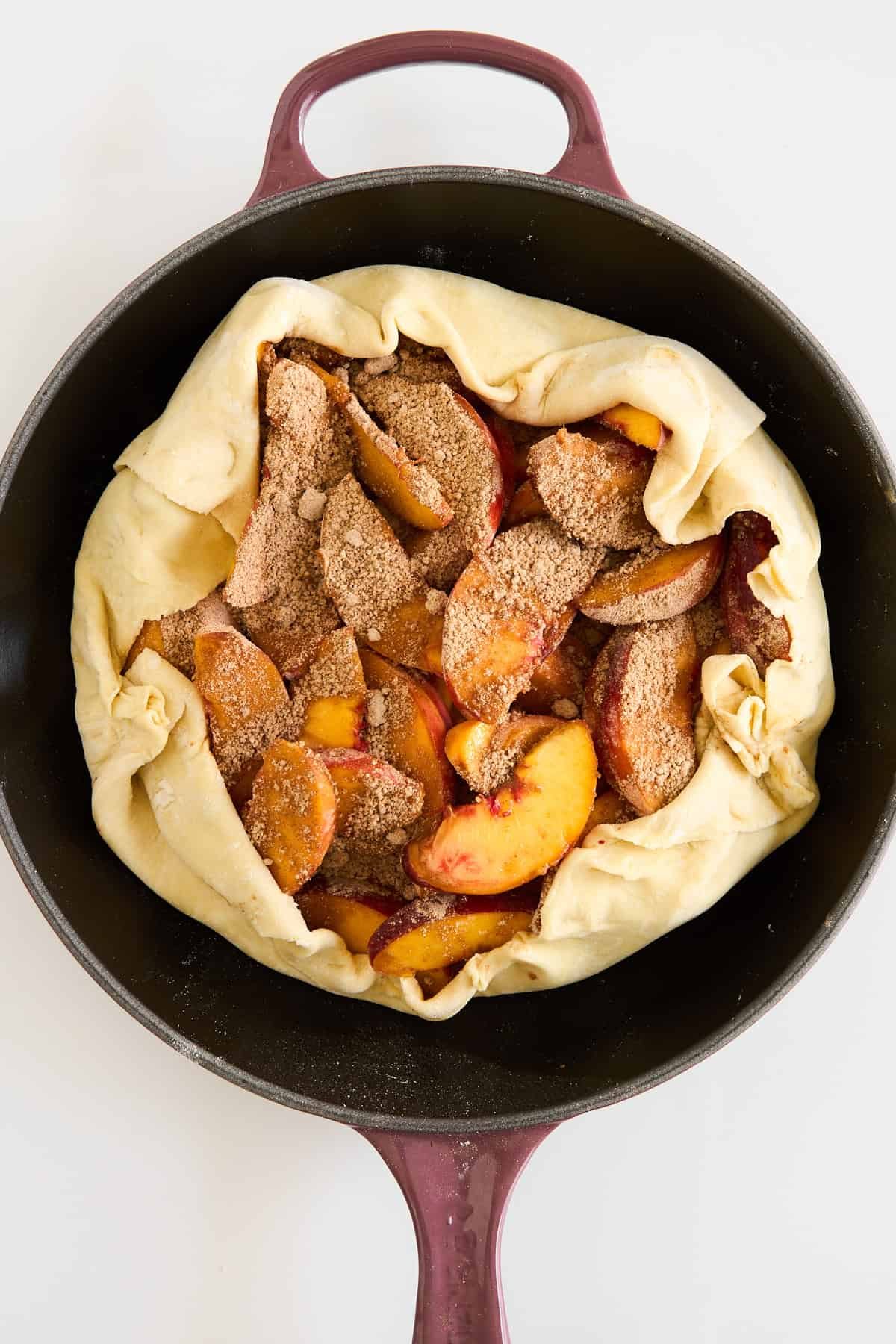 A cast iron skillet holds an unbaked rustic peach galette with folded pastry edges, filled with sliced peaches coated in brown sugar and spices.