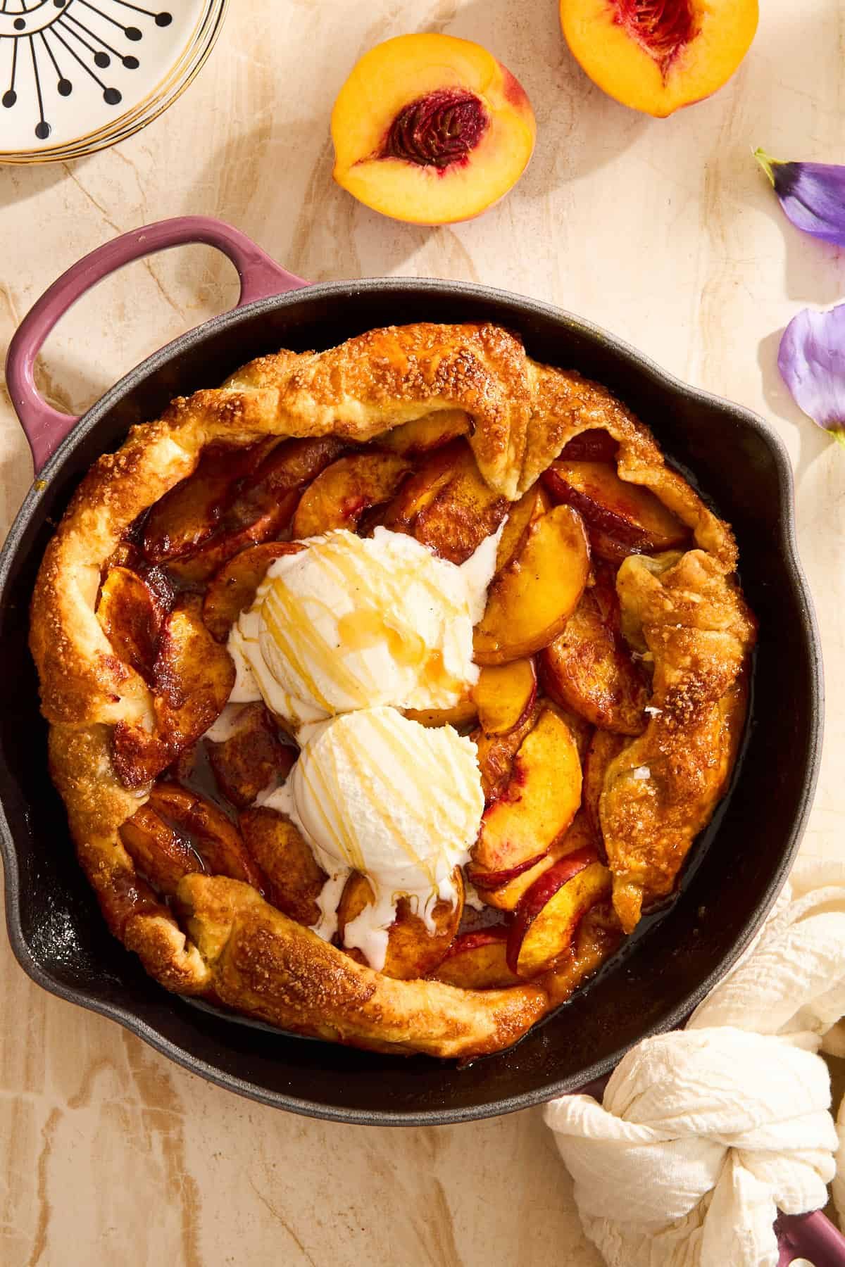 A peach galette with a golden crust sits in a black skillet, topped with two scoops of vanilla ice cream and a drizzle of honey. Slices of fresh peach and a floral napkin are nearby on a light-colored surface.
