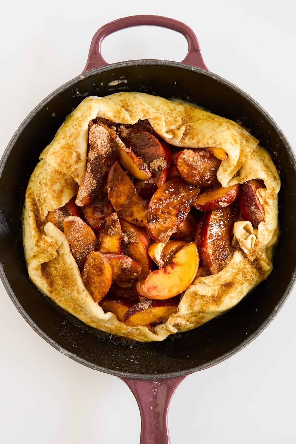 A rustic peach galette with golden crust and sliced peaches, sprinkled with cinnamon and sugar, sits in a round, dark baking dish with handles on a white background.