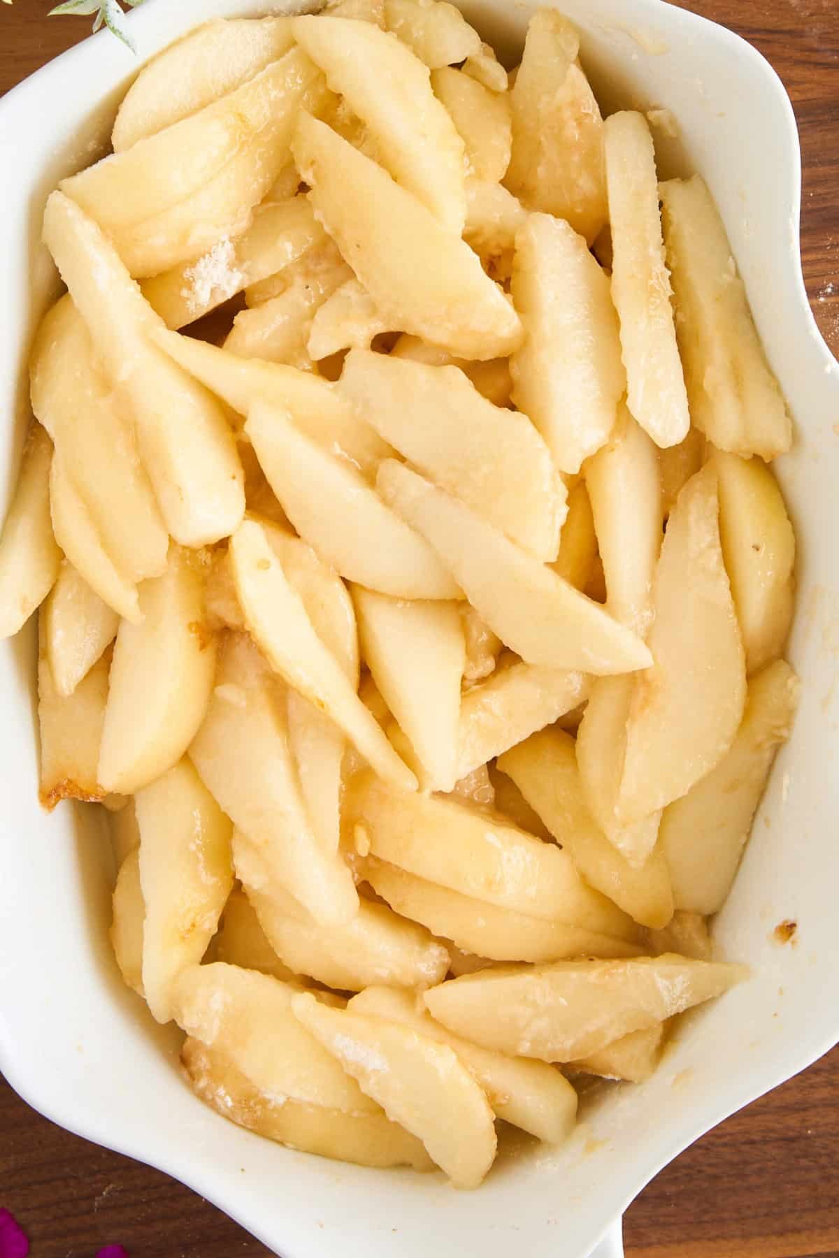 Sliced fresh pears coated with sugar and arranged in a white ceramic baking dish, ready for baking. The pears have a glossy appearance and are evenly distributed in the dish.