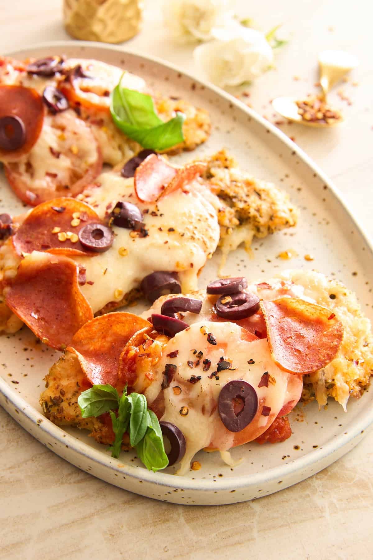 A plate of baked chicken topped with melted cheese, pepperoni slices, black olives, red pepper flakes, and fresh basil, served on a speckled oval dish.