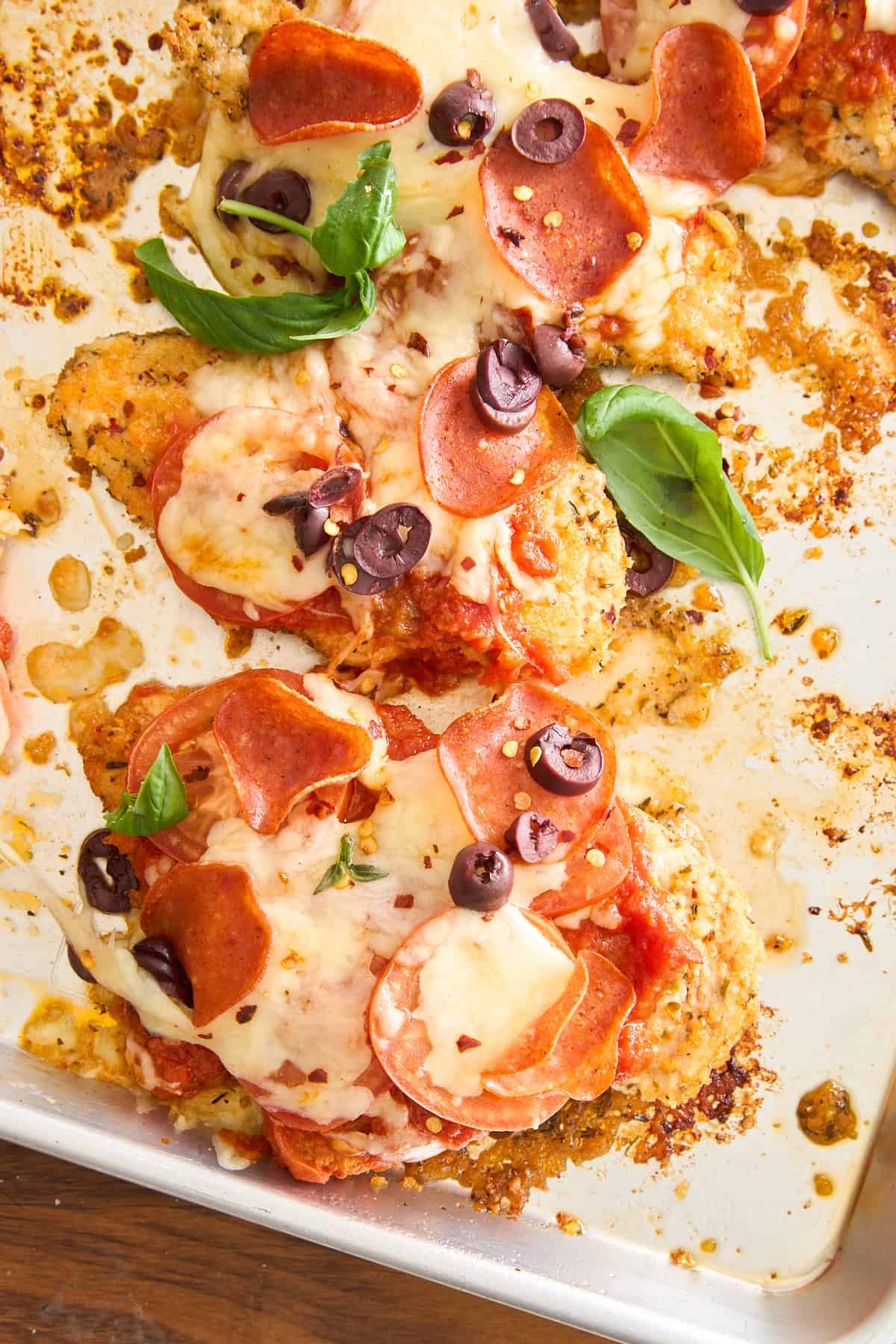Baked chicken breasts topped with melted cheese, pepperoni slices, black olives, tomato sauce, and fresh basil leaves on a sheet pan.