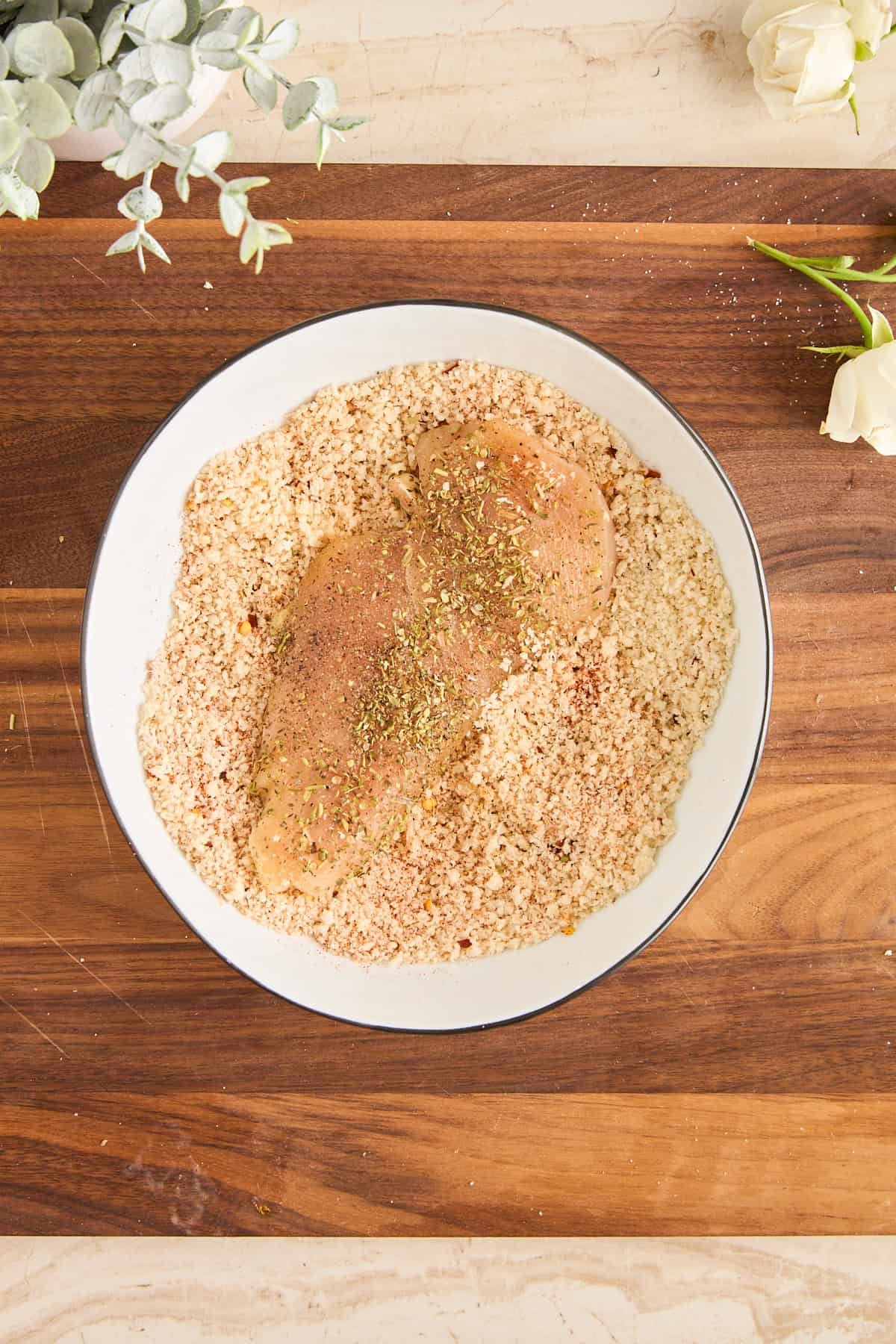 A raw, seasoned chicken breast rests in a bowl of breadcrumbs on a wooden cutting board. Greenery and white flowers decorate the edges of the scene.