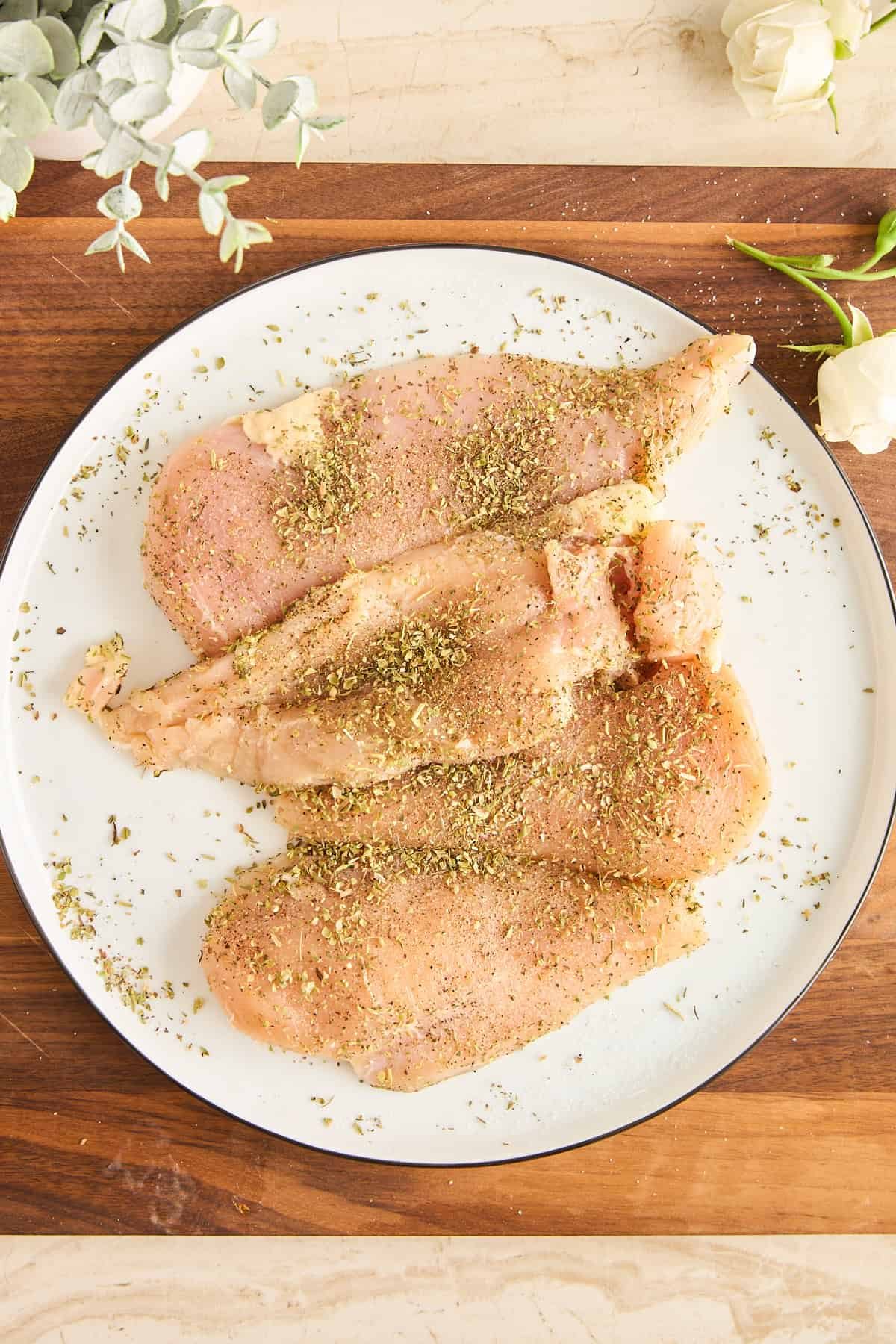 Four raw, seasoned chicken breasts are arranged on a white plate, sprinkled with herbs and spices. The plate sits on a wooden cutting board, with white flowers and green leaves nearby.