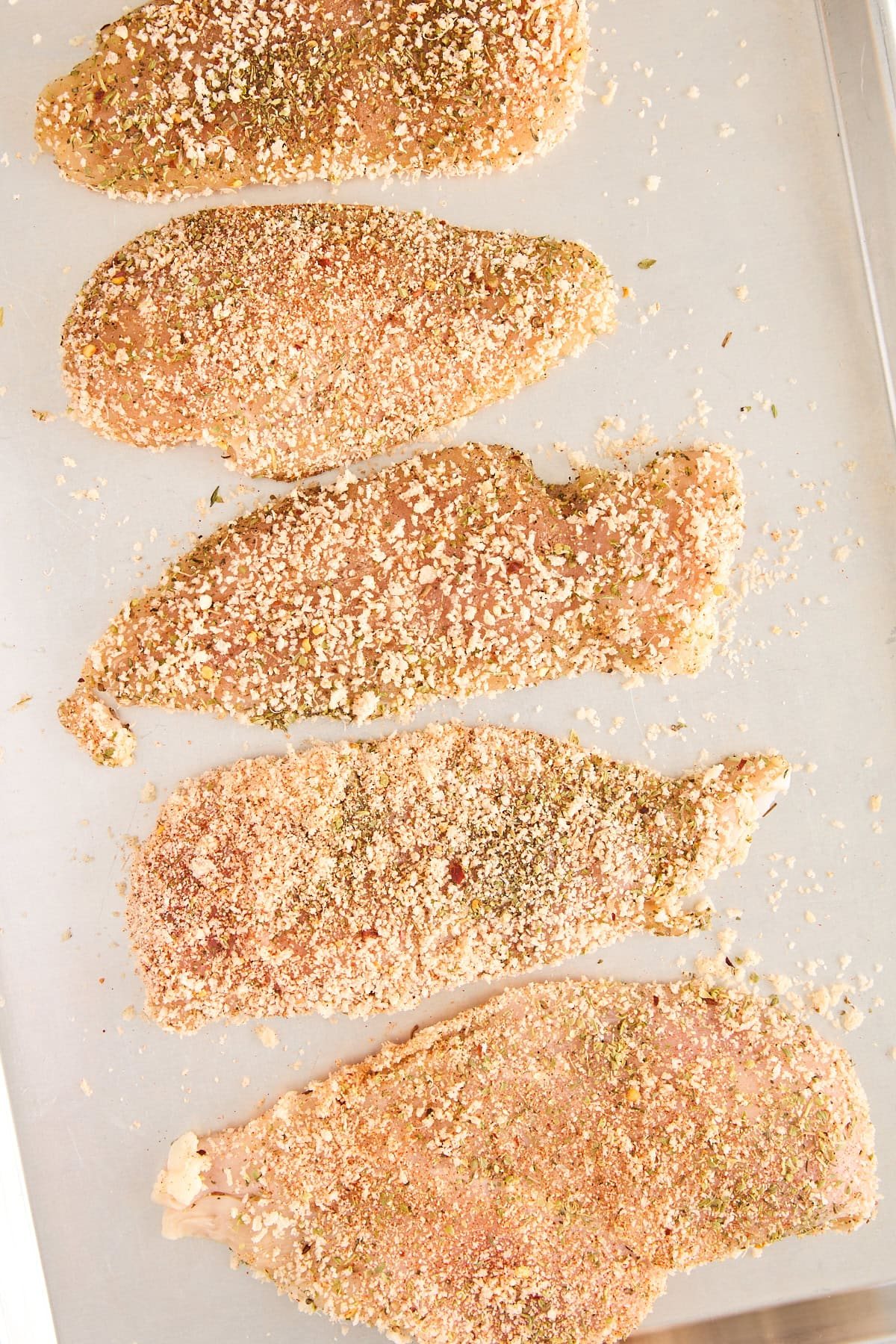 Five raw, breaded chicken cutlets are arranged in a row on a metal baking sheet, coated with herbs and breadcrumbs, ready to be cooked.
