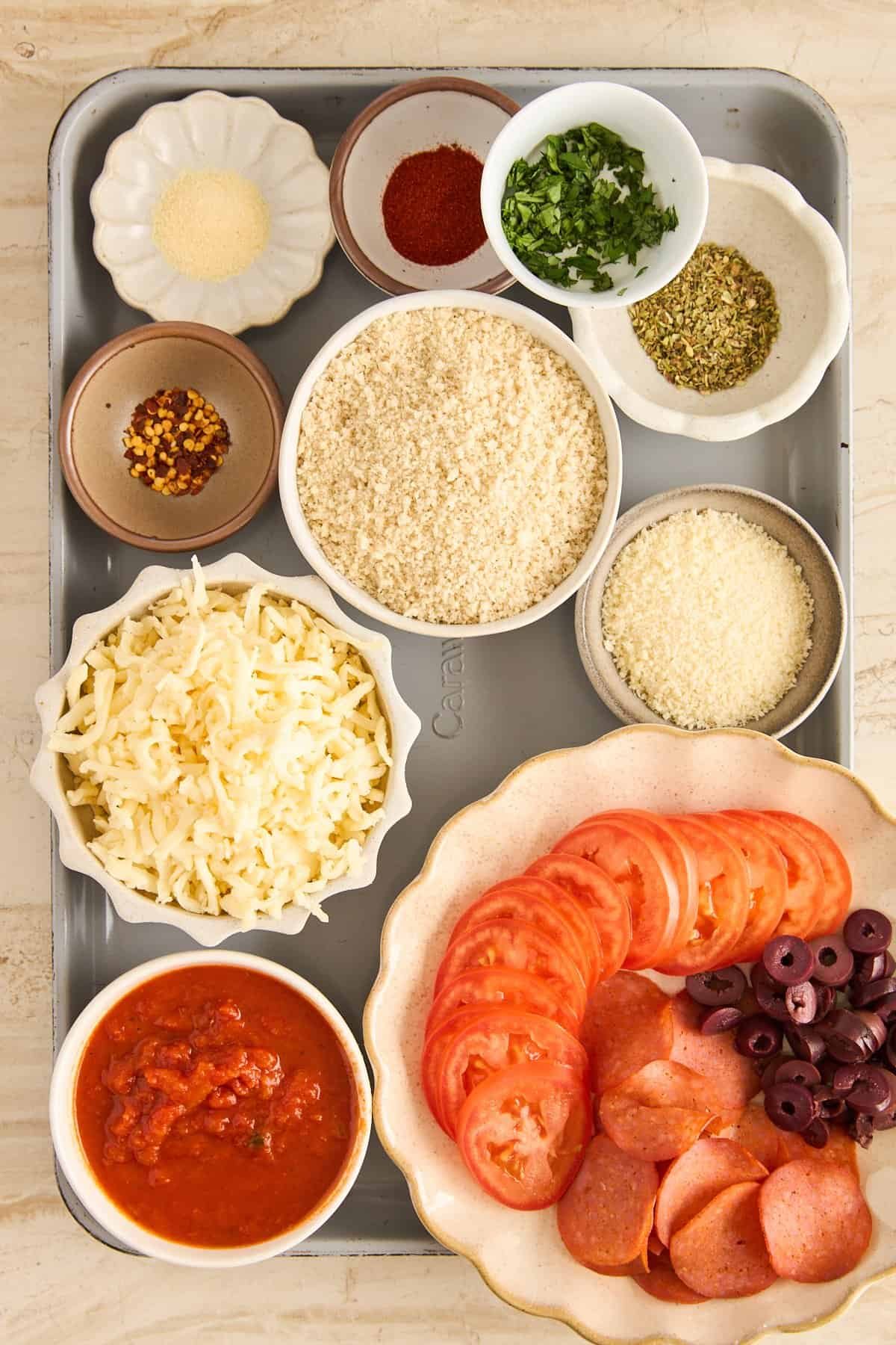 A tray with bowls of shredded cheese, tomato slices, sliced pepperoni, black olives, marinara sauce, breadcrumbs, grated parmesan, and small bowls of various spices and herbs.