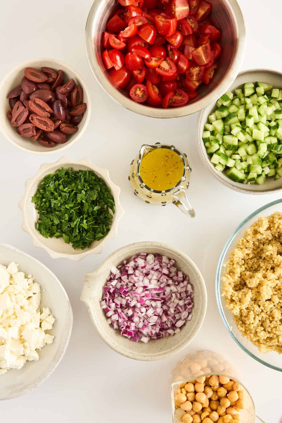Bowls filled with chopped tomatoes, cucumbers, red onions, parsley, feta cheese, Kalamata olives, cooked quinoa, chickpeas, and a small pitcher of yellow salad dressing arranged on a white surface.