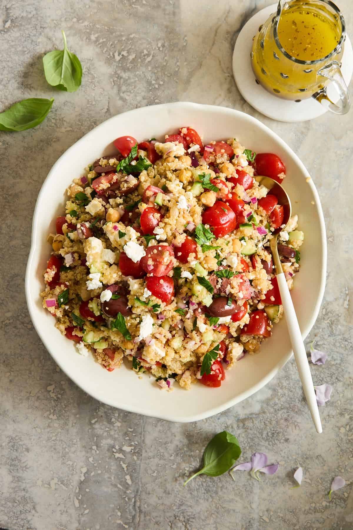 A white bowl filled with a colorful salad of couscous, cherry tomatoes, feta cheese, red onion, and fresh greens, with a white spoon. A small jar of yellow dressing sits nearby on a marble surface.