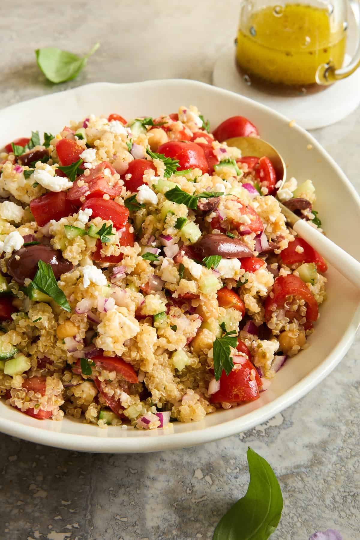 A bowl of quinoa salad with chopped tomatoes, cucumbers, red onions, kalamata olives, parsley, and crumbled feta cheese, with a spoon and a small jar of dressing in the background.