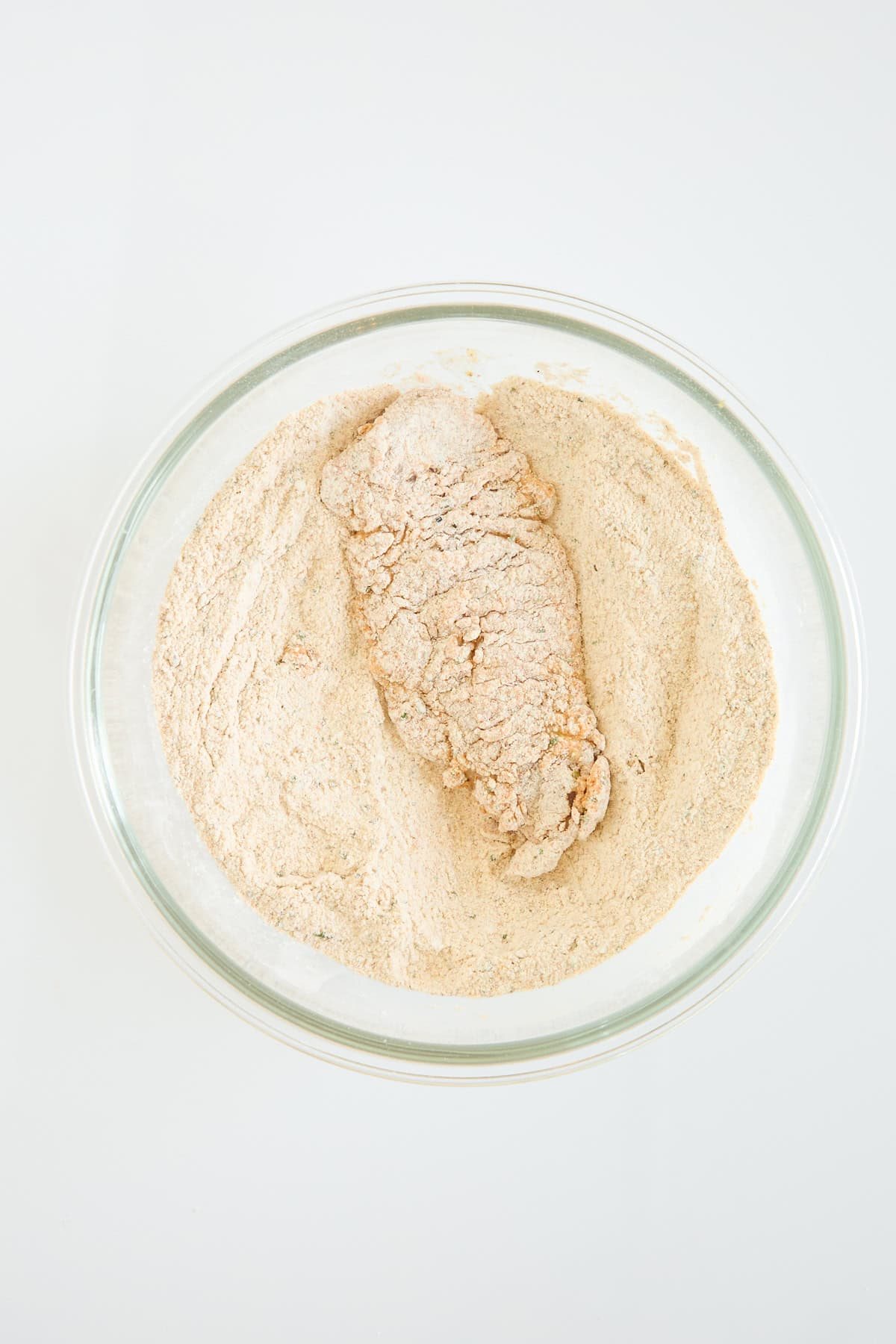 A piece of food, likely chicken, coated in flour and seasonings sits in a clear glass bowl filled with more flour mixture on a white surface.