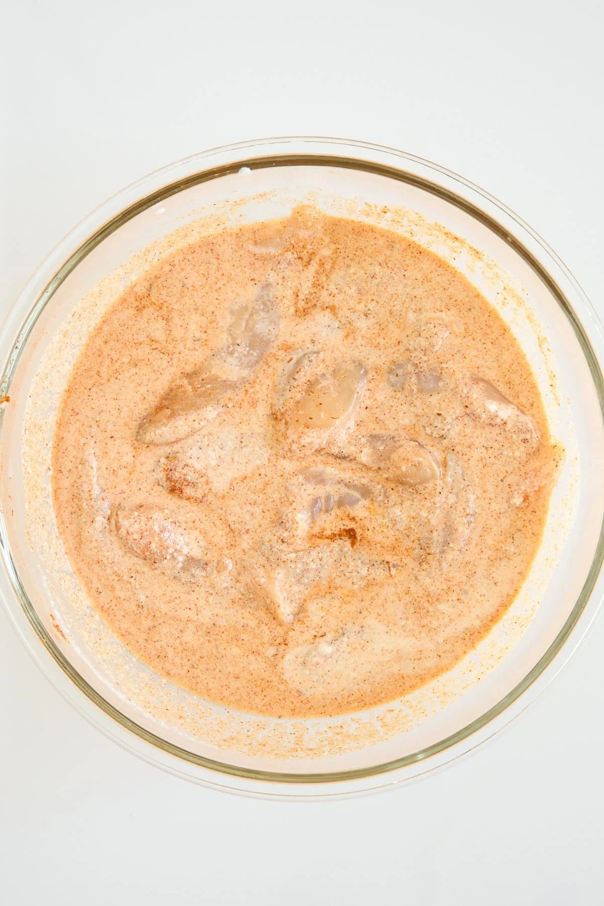 A clear glass bowl filled with pieces of chicken marinating in a creamy, orange-colored spice mixture, set against a plain white background.