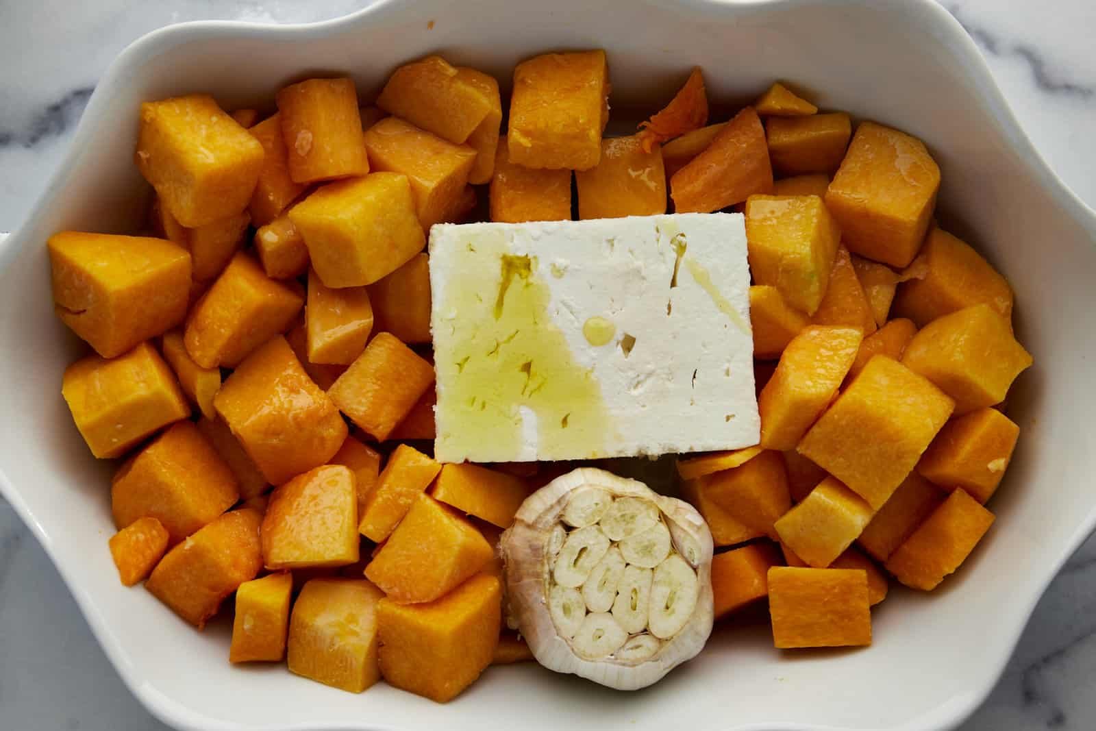 Butternut squash cubes, a block of feta, and a head of garlic topped with oil in a baking dish.