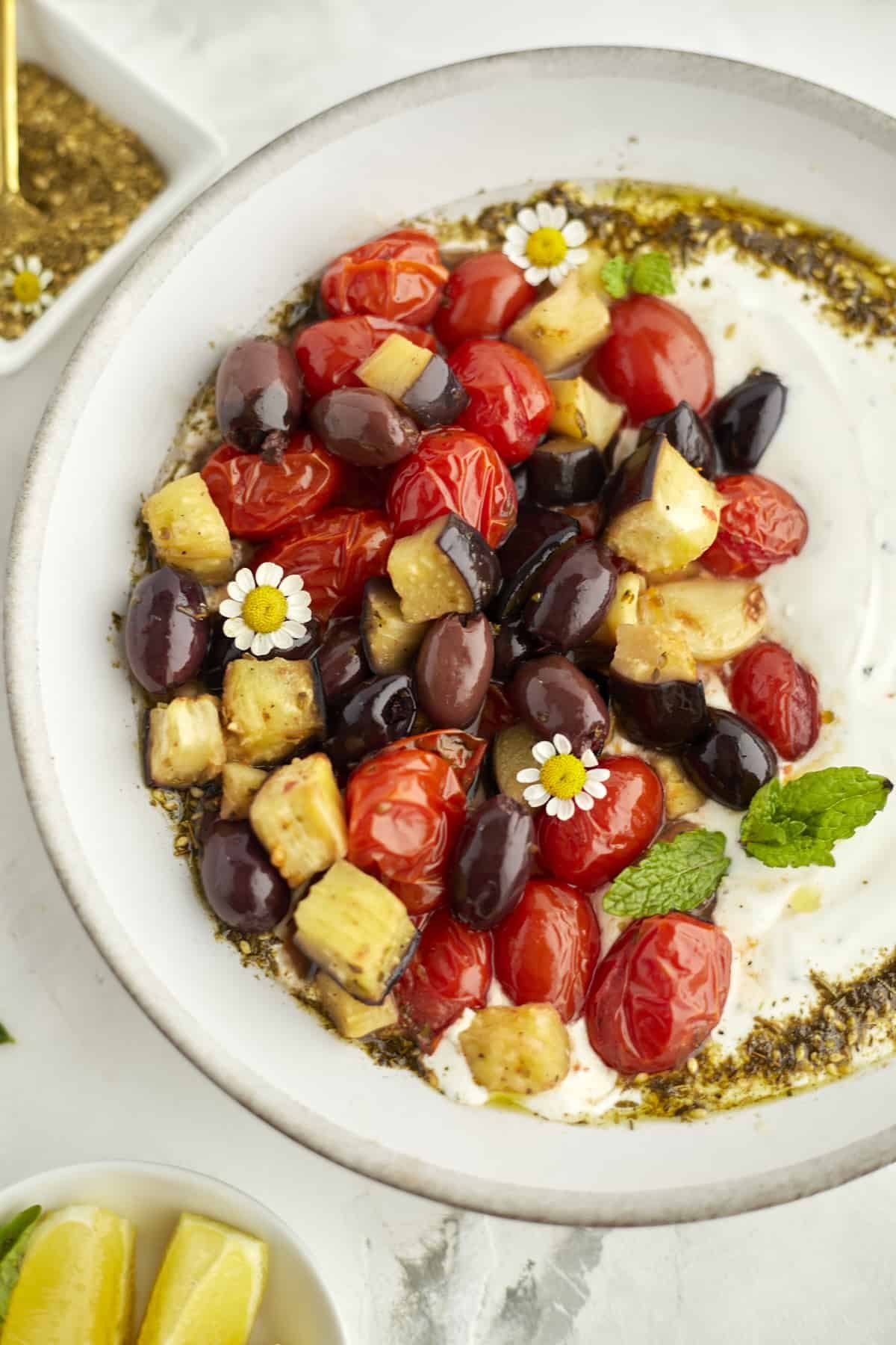 Plated eggplant, tomatoes and olives over creamy yogurt dip. Garnished with zaatar and fresh mint