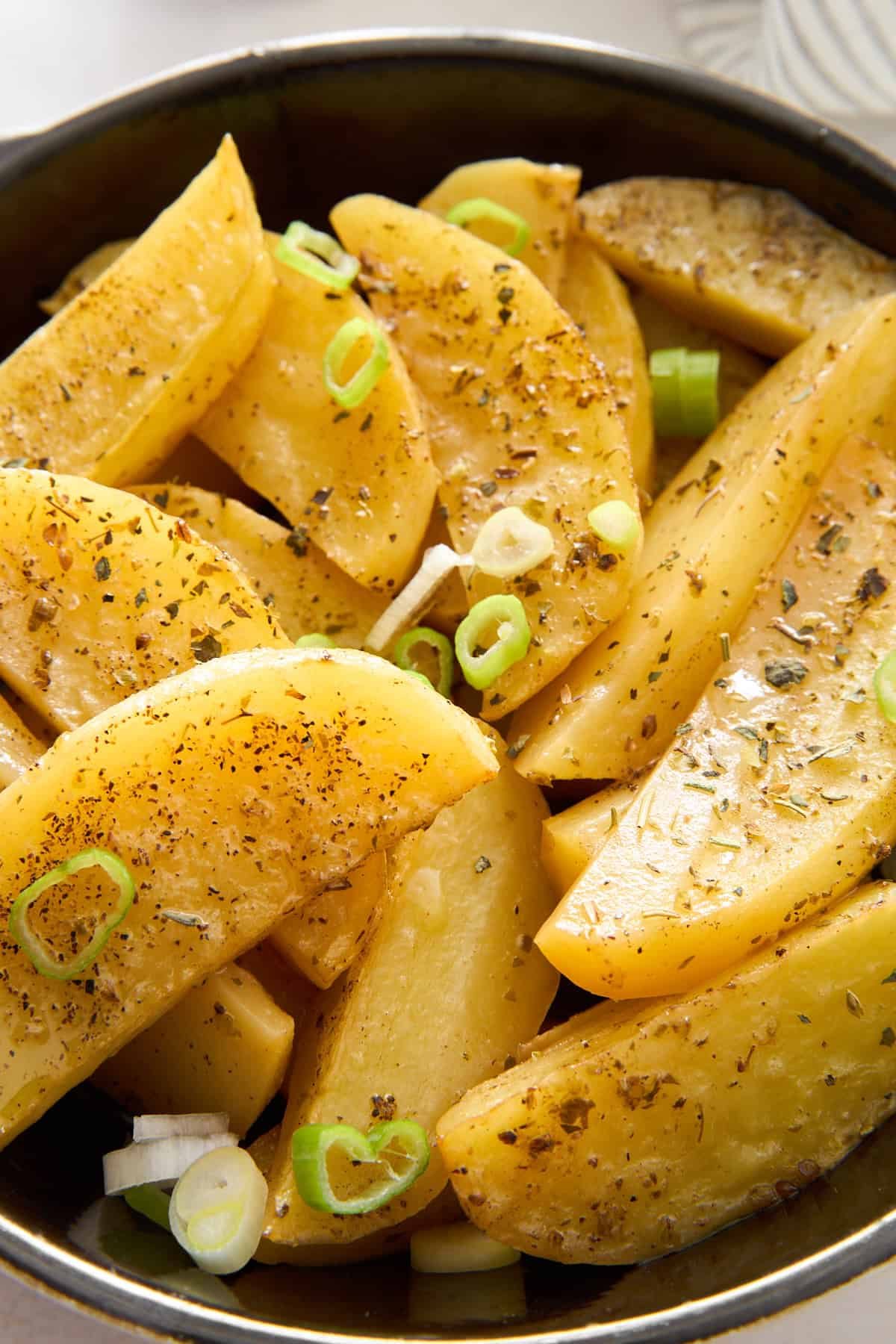 A bowl of seasoned potato wedges garnished with sliced green onions and herbs, showing a close-up of the golden, baked potatoes with visible spices on their surface.